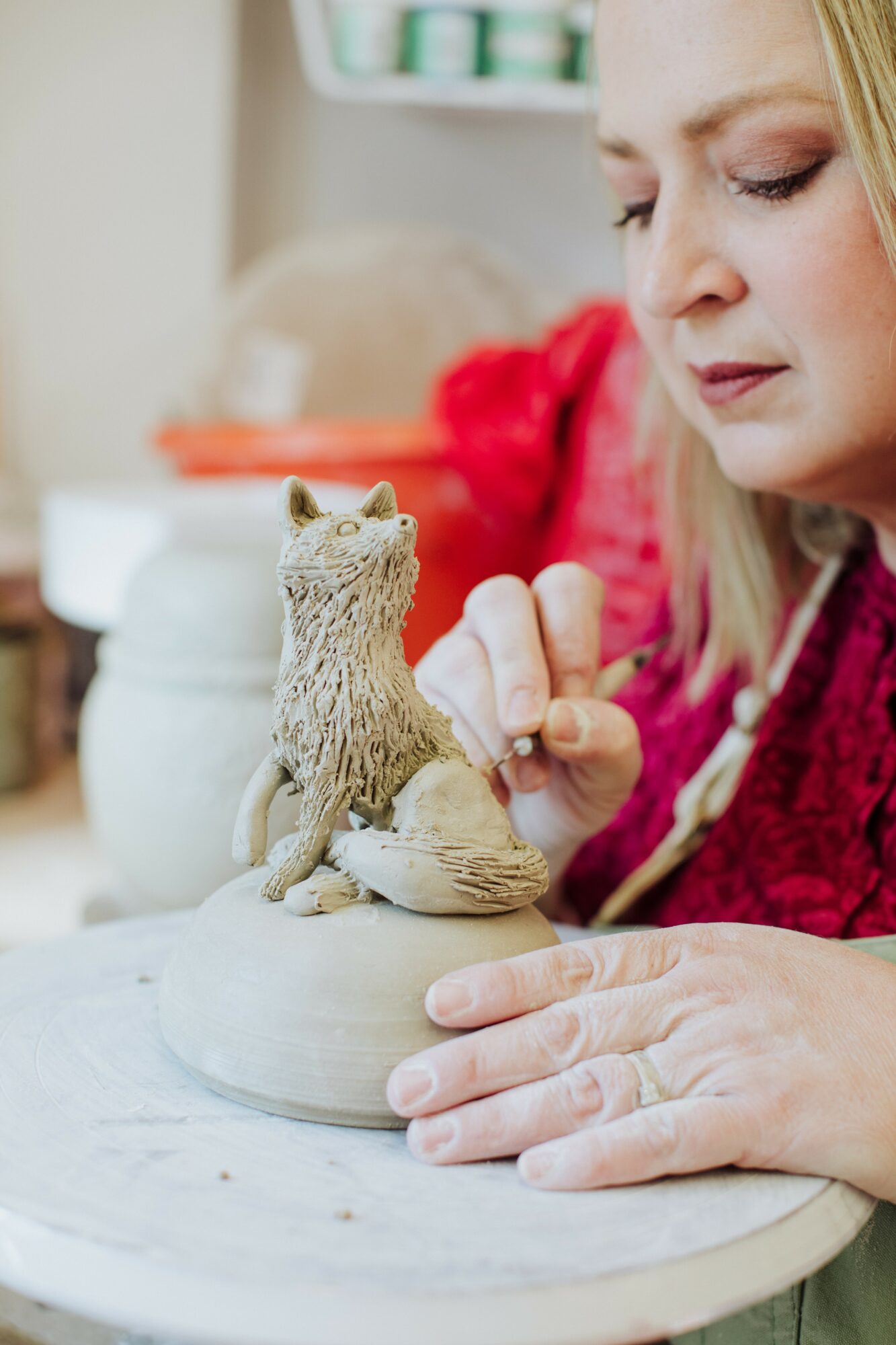 Woman painting a small animal sculpture on a round base, with a blurred background, wearing a red garment.