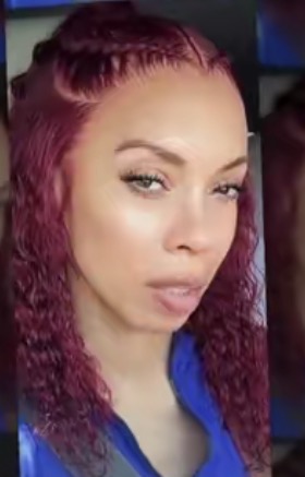 Close-up of a woman with red, curly hair and makeup, wearing a blue top, looking at the camera.