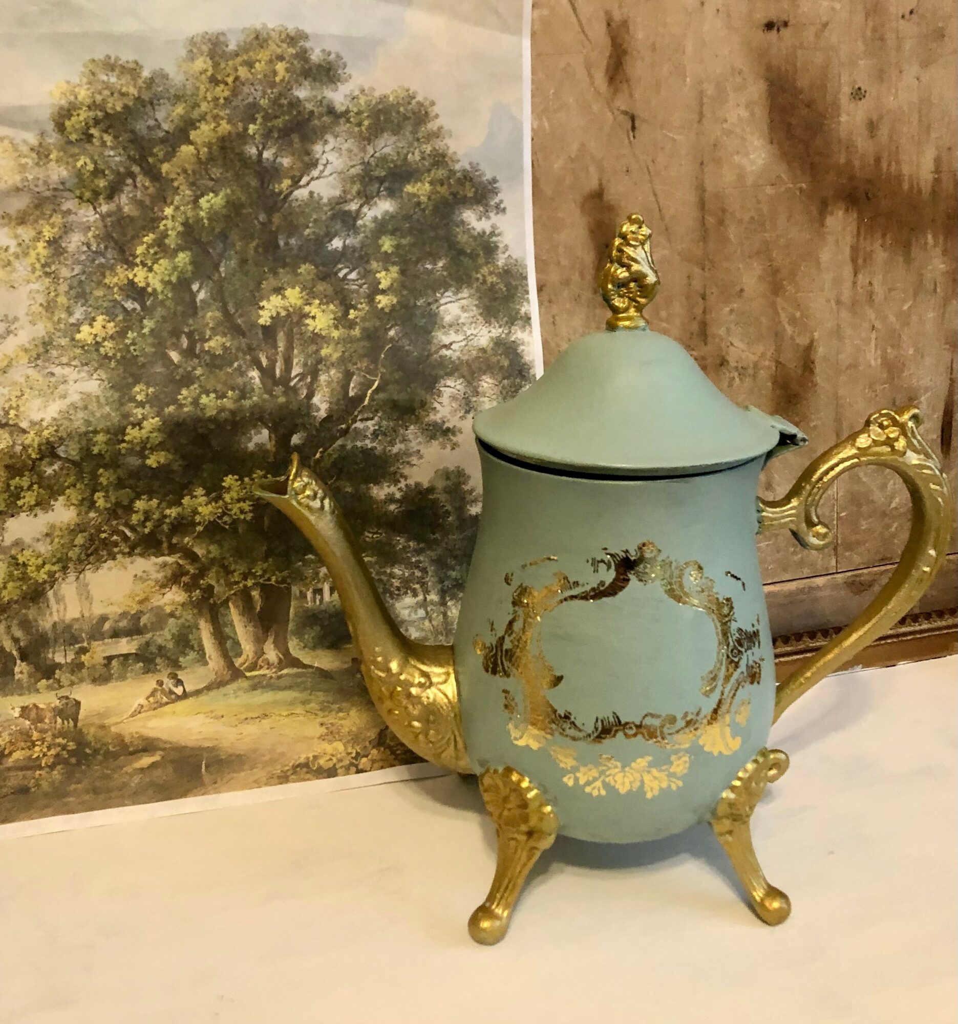 Decorative teapot with gold accents and ornate legs, placed in front of a landscape painting of a large tree.