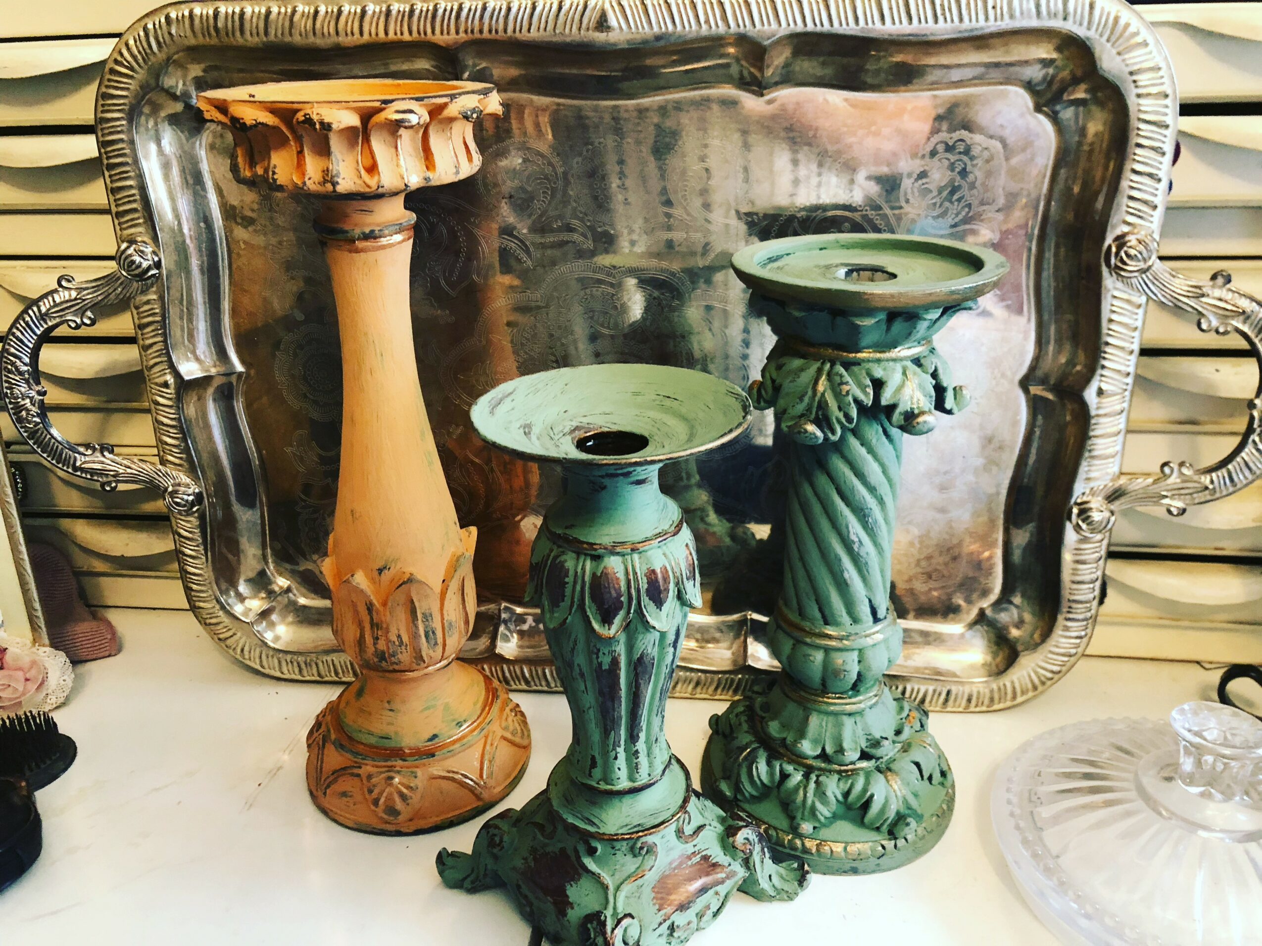 Three decorative candle holders of varying heights and colors in front of a silver tray, on a surface with other items.
