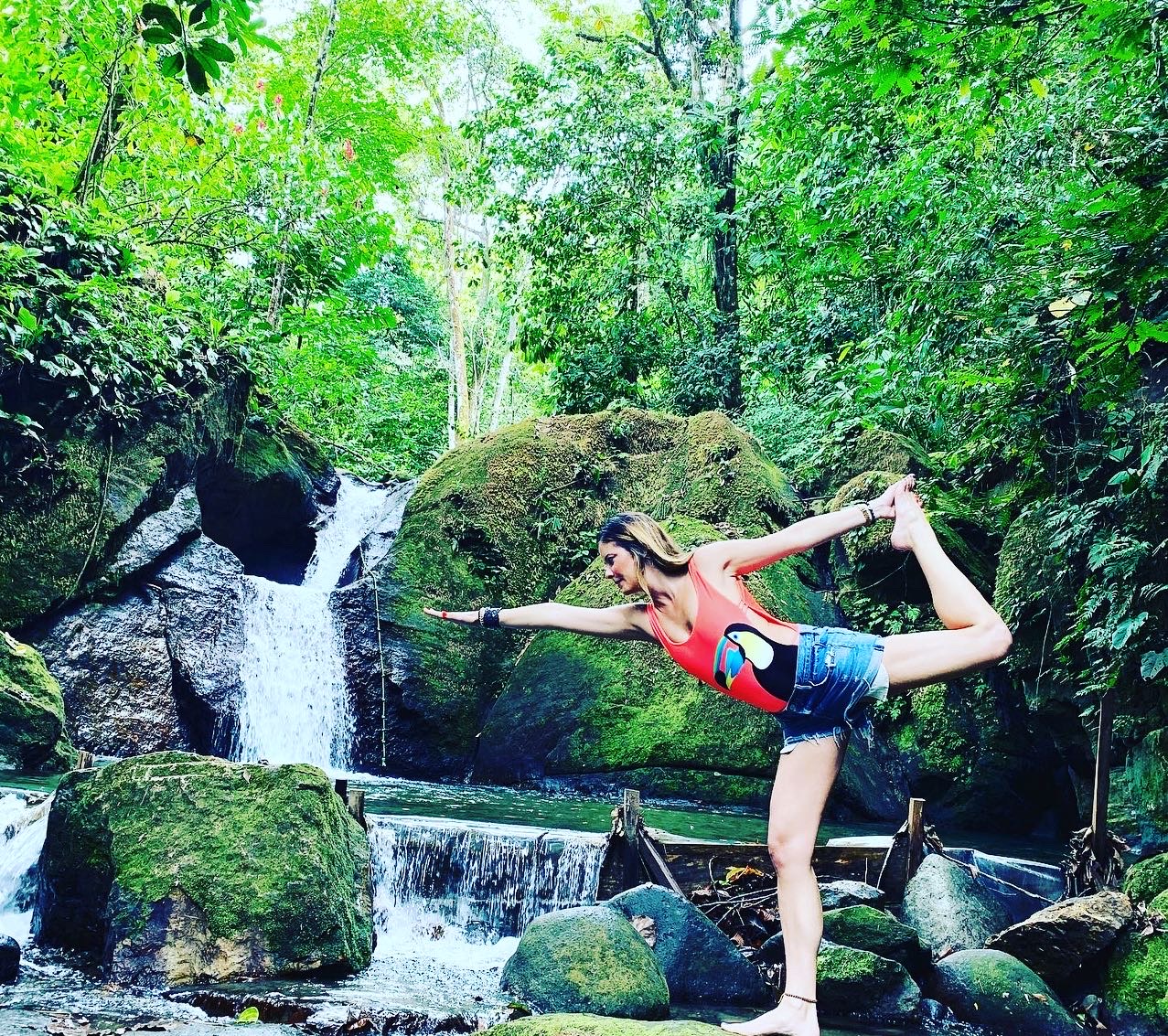 Person in a yoga pose balancing on one leg with arms extended, in a lush forest near a waterfall.