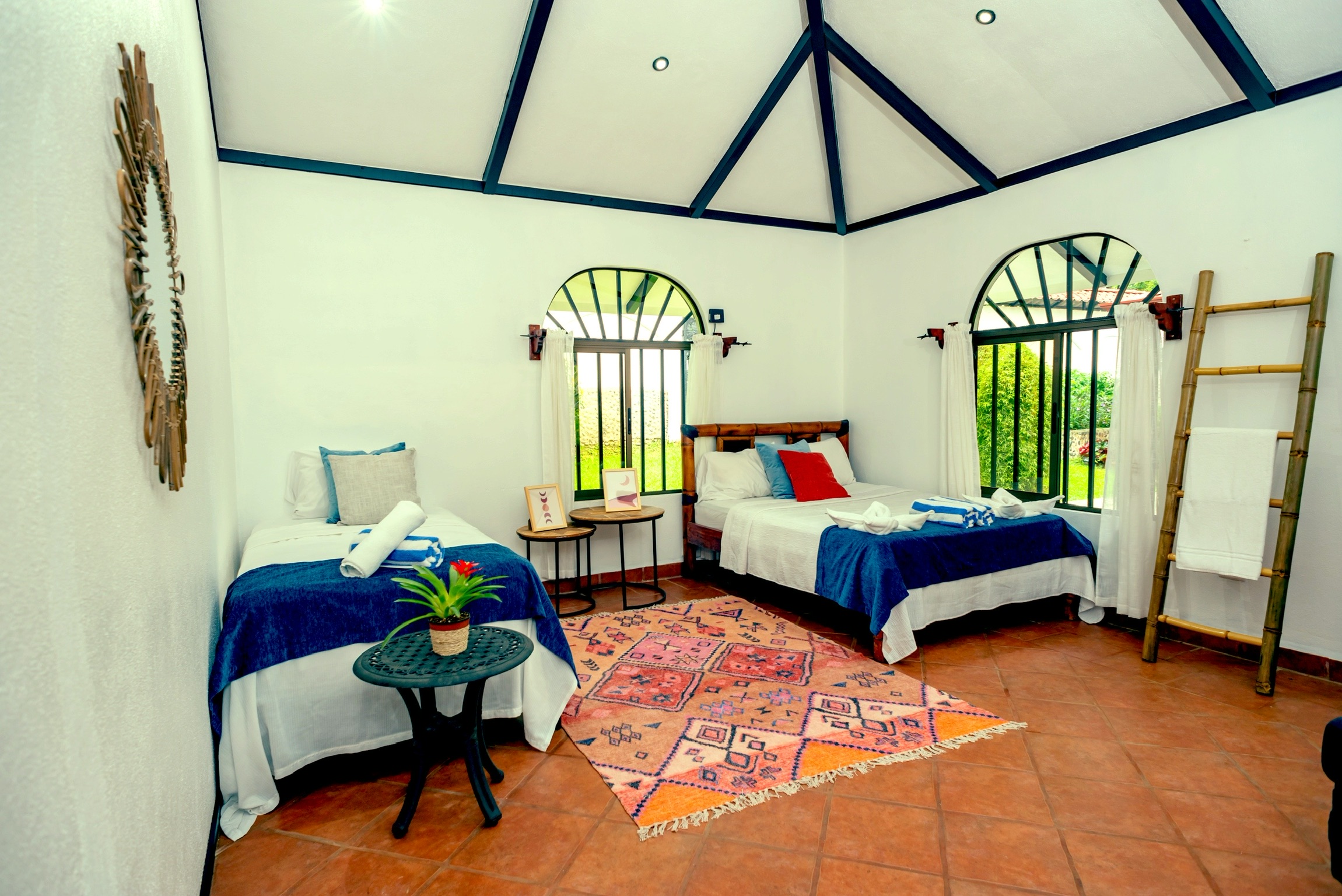 Bedroom with two beds, arched windows, a colorful rug, and a ladder, with white walls and a high ceiling.