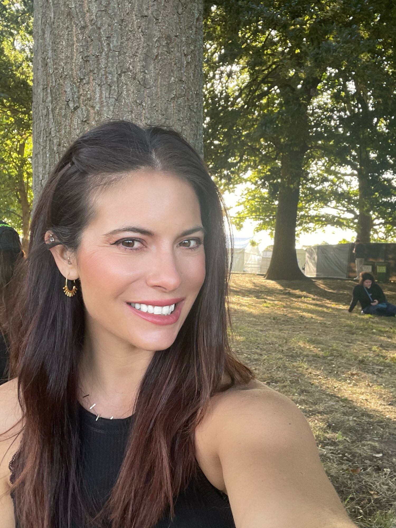 Young woman with long dark hair smiling outdoors near a tree, sunlight filtering through leaves, people sitting in background.
