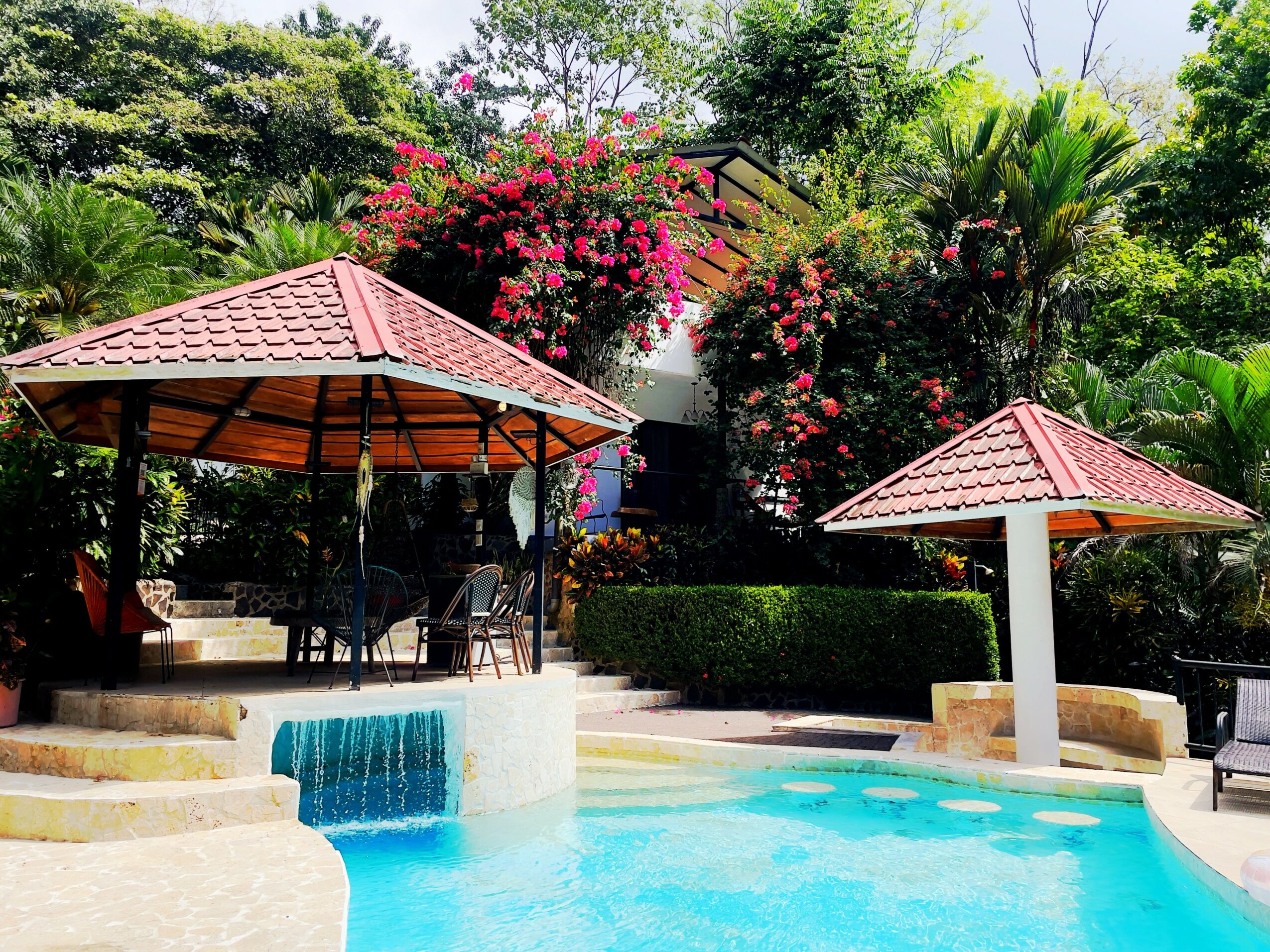 Swimming pool with two red-roofed gazebos surrounded by lush green trees and flowering bushes.