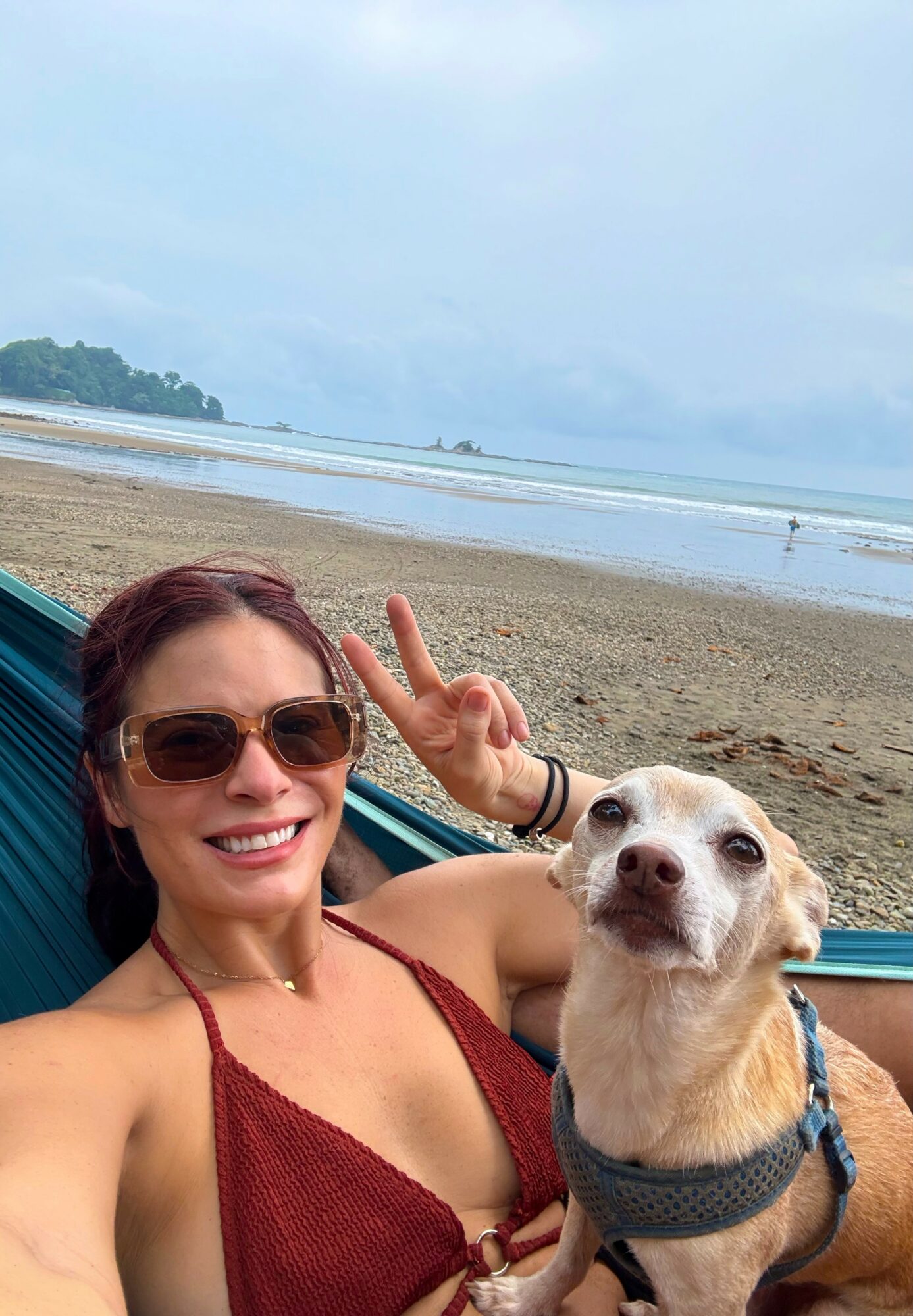 Woman with sunglasses smiling and making a peace sign, lying in a hammock with a dog on a beach.