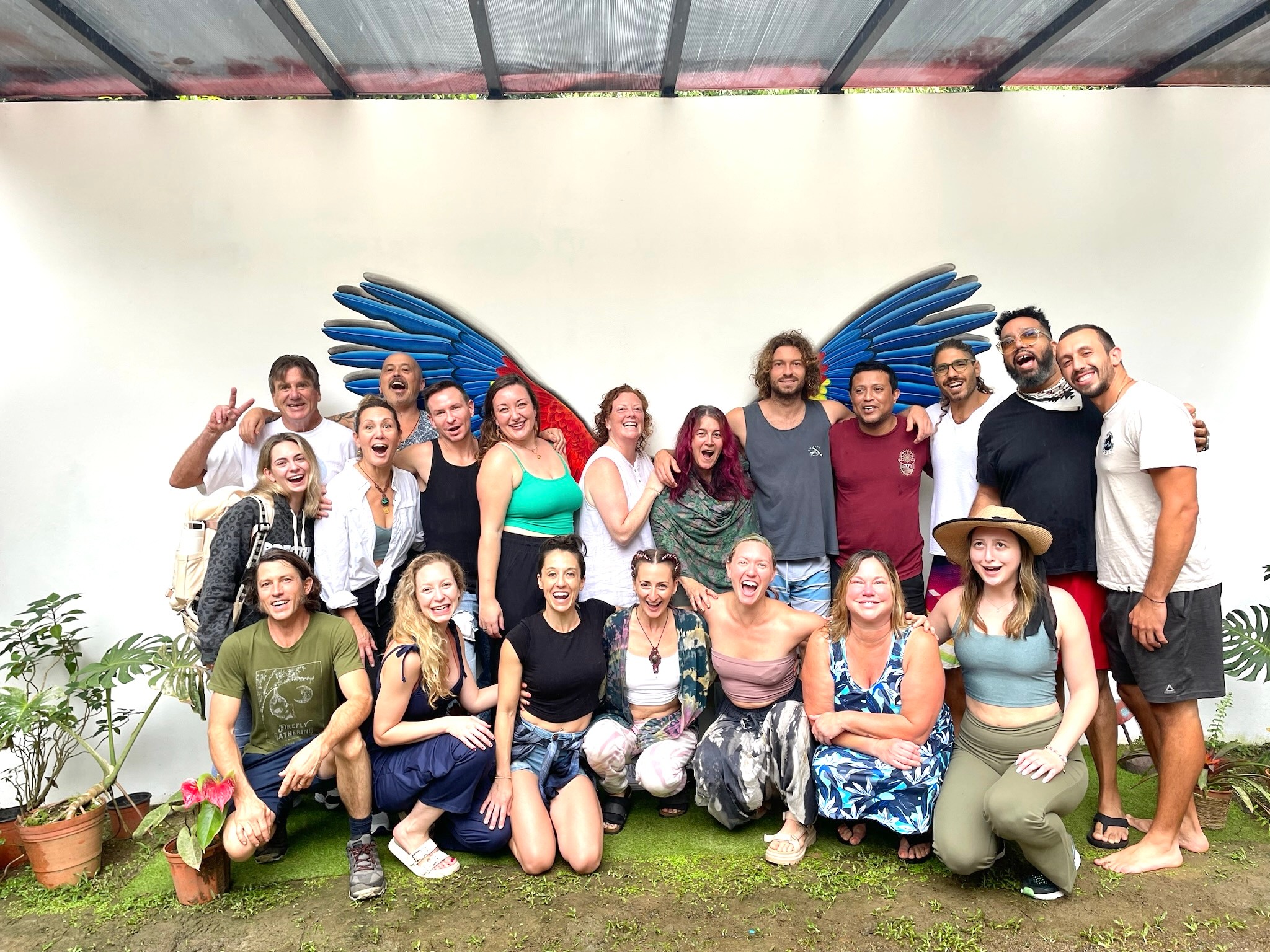 Group of 17 people outdoors, some with wings, smiling and posing together, with plants and a white wall background.