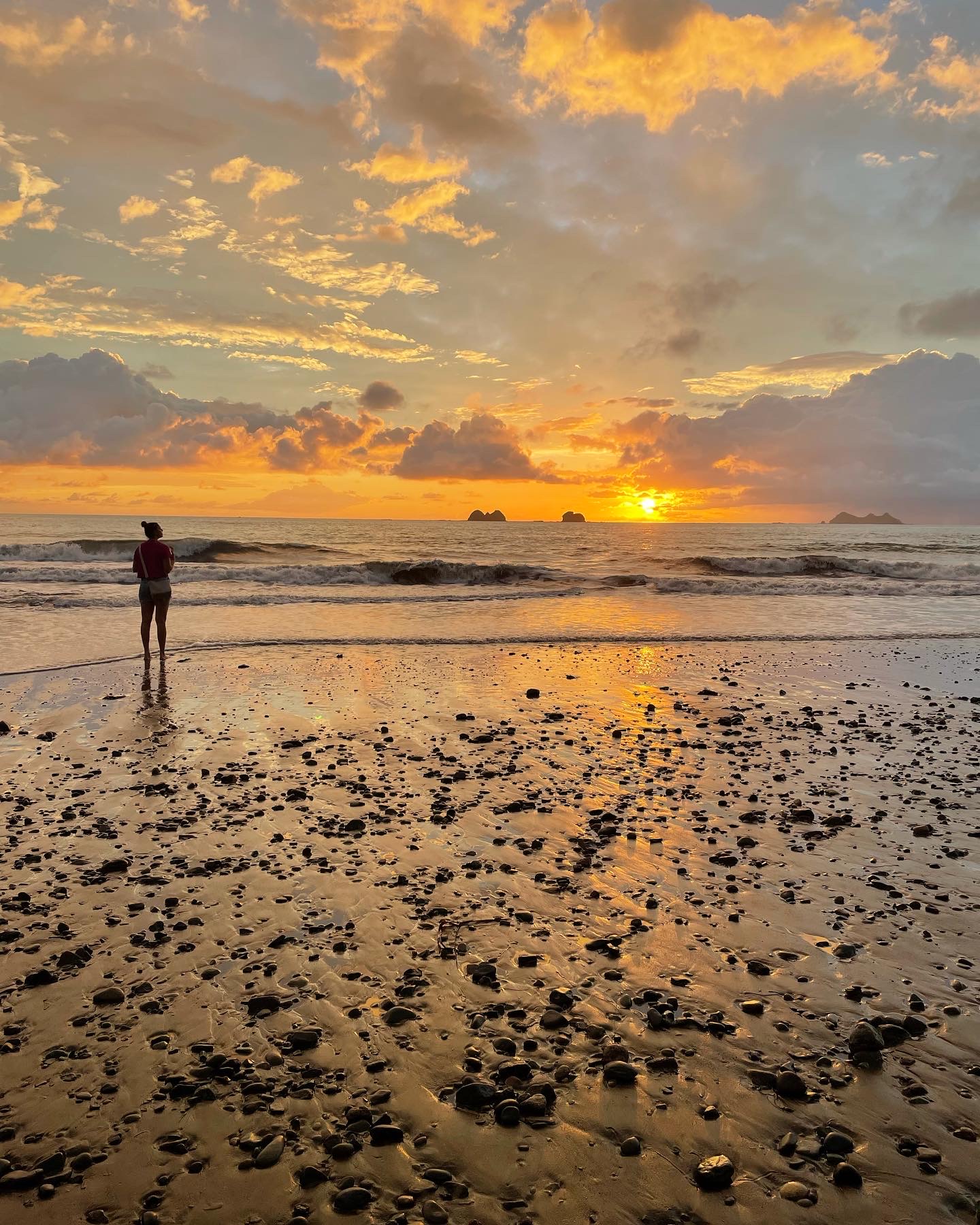 Person standing on a beach during sunset with clouds and ocean waves, sandy shore with rocks, warm colors in sky.