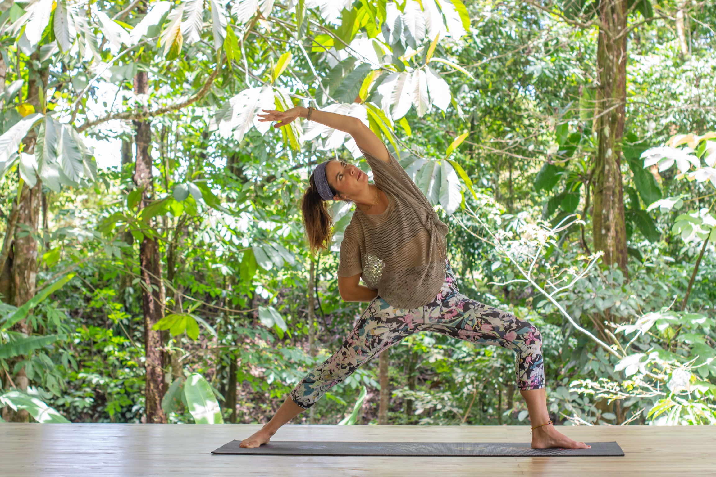 Person practicing yoga outdoors in a forest, performing a wide-legged stretch with one arm raised overhead.