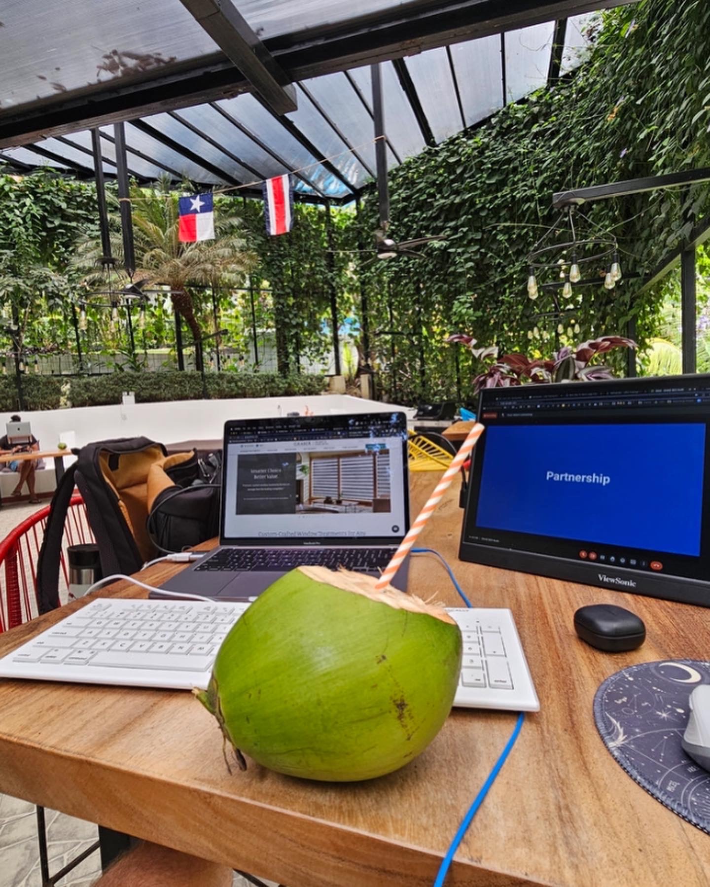 Green coconut on a wooden table with laptops and a mouse, outdoor setting with flags and greenery in the background.