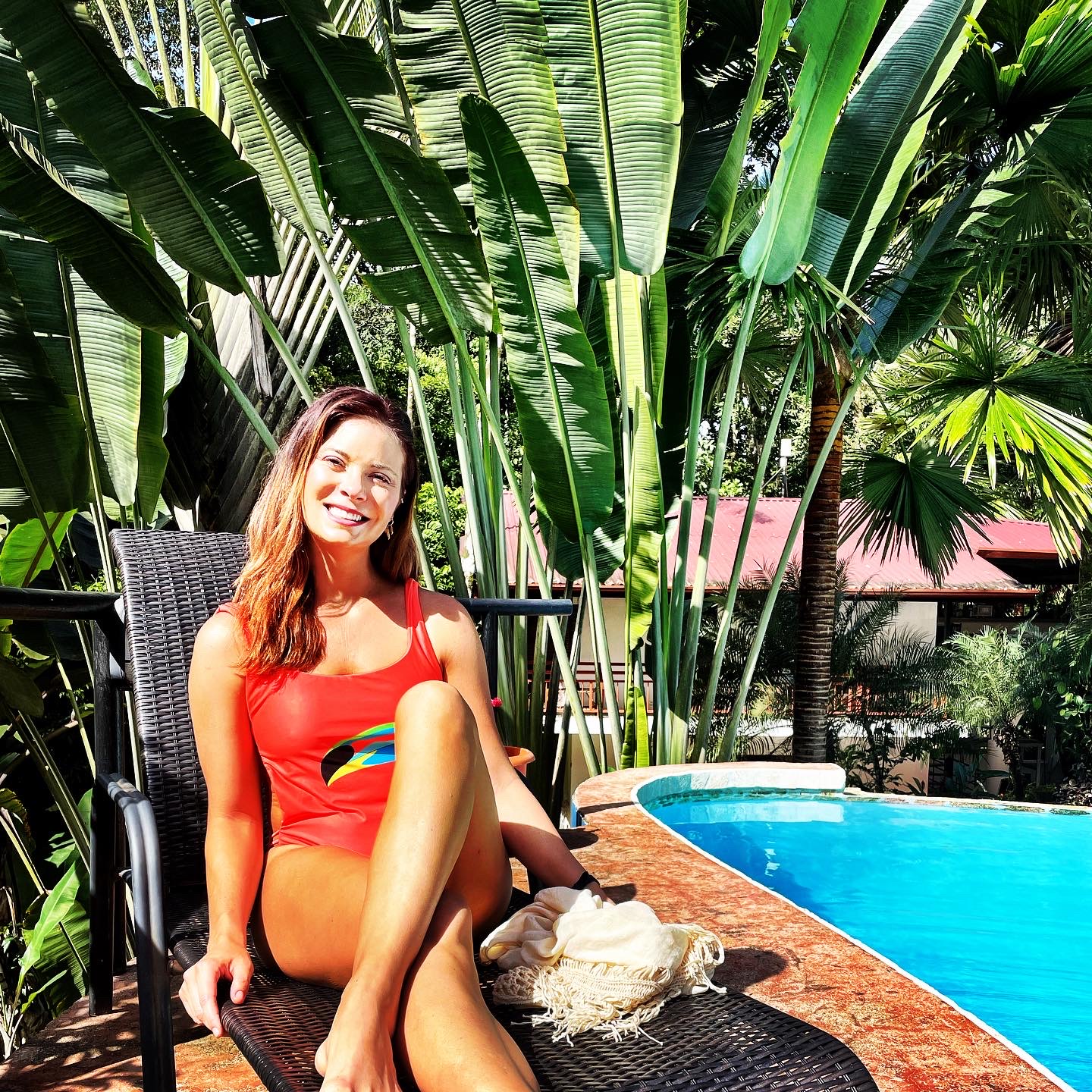 Smiling woman in red swimsuit sitting by a pool surrounded by large green tropical plants.