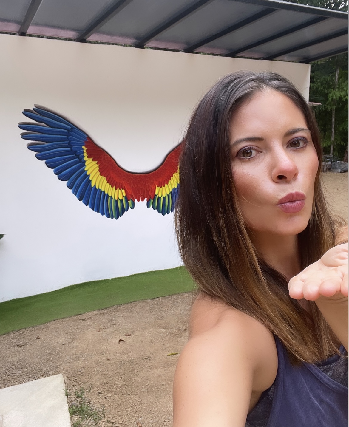 Woman with long brown hair blowing a kiss in front of a colorful wing mural on a white wall, outdoors.