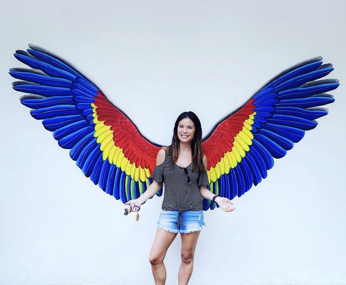 Woman standing in front of colorful wing mural, smiling, arms outstretched, white background, casual clothing, shorts, long dark hair