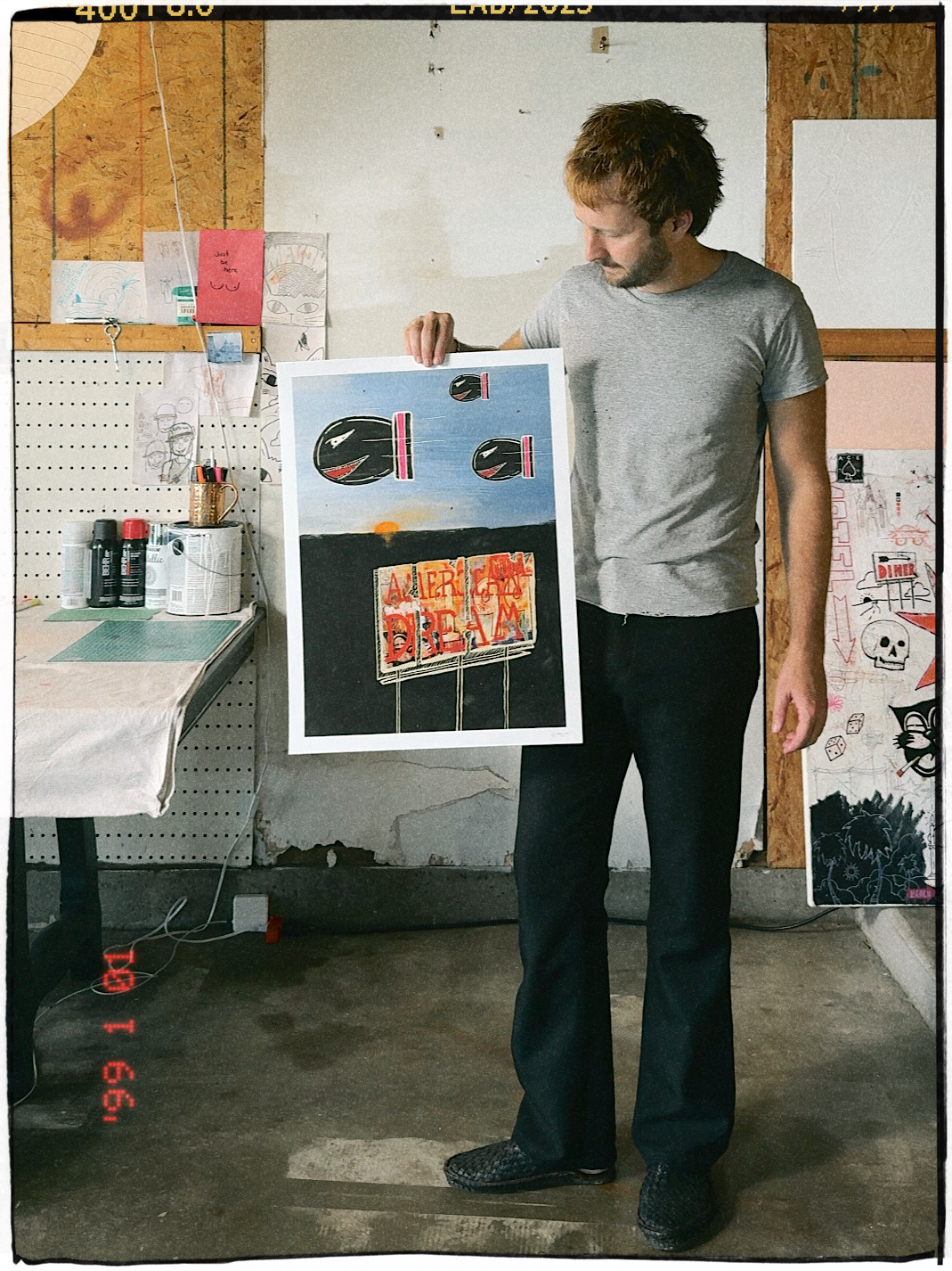Man holding artwork in a workshop with pegboard wall and supplies, looking at the art piece.