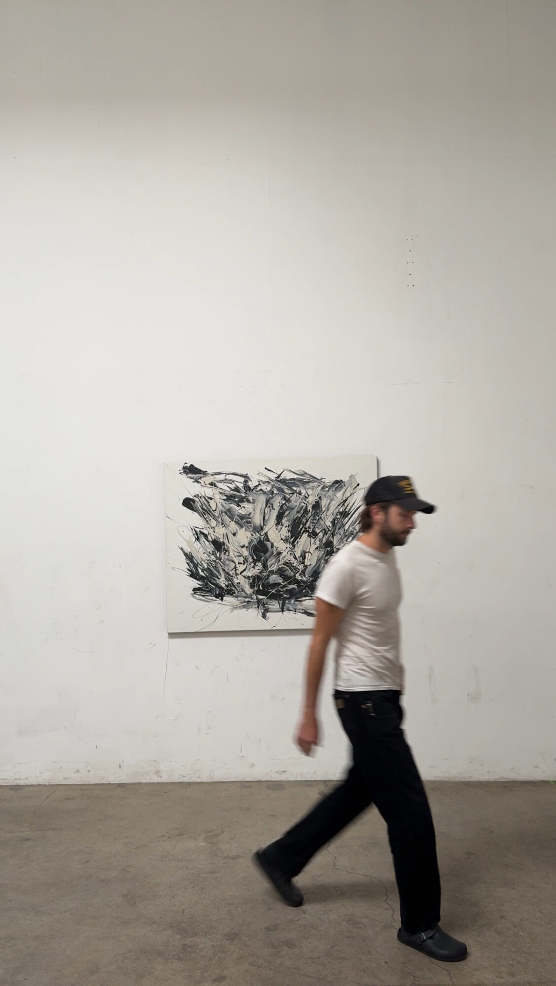 Person walking past abstract black and white artwork on a white wall, wearing a cap, white t-shirt, and dark pants.