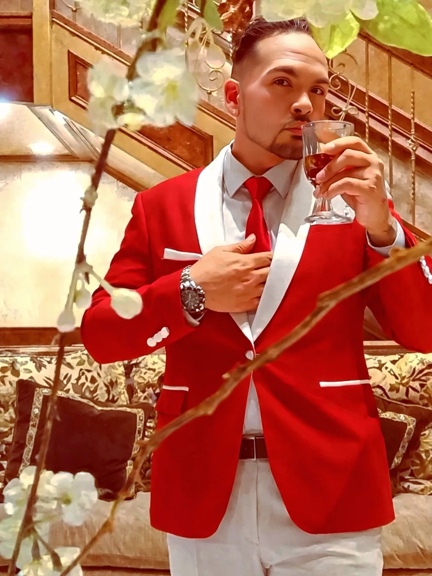 Man in red blazer and white shirt holding a glass of wine indoors, with floral decorations and a staircase in background.