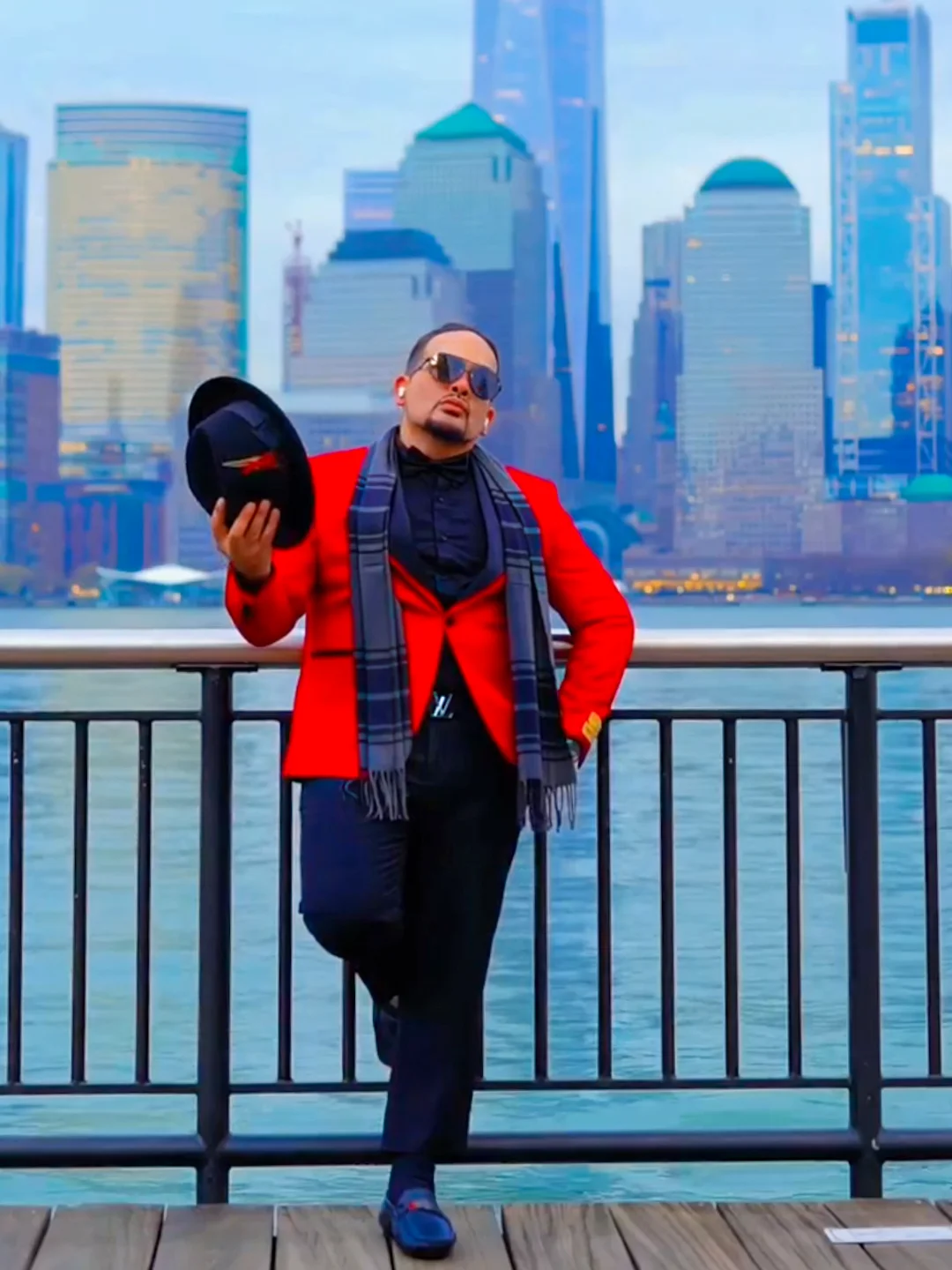 Man in red jacket and sunglasses standing by a railing with city skyline in background.