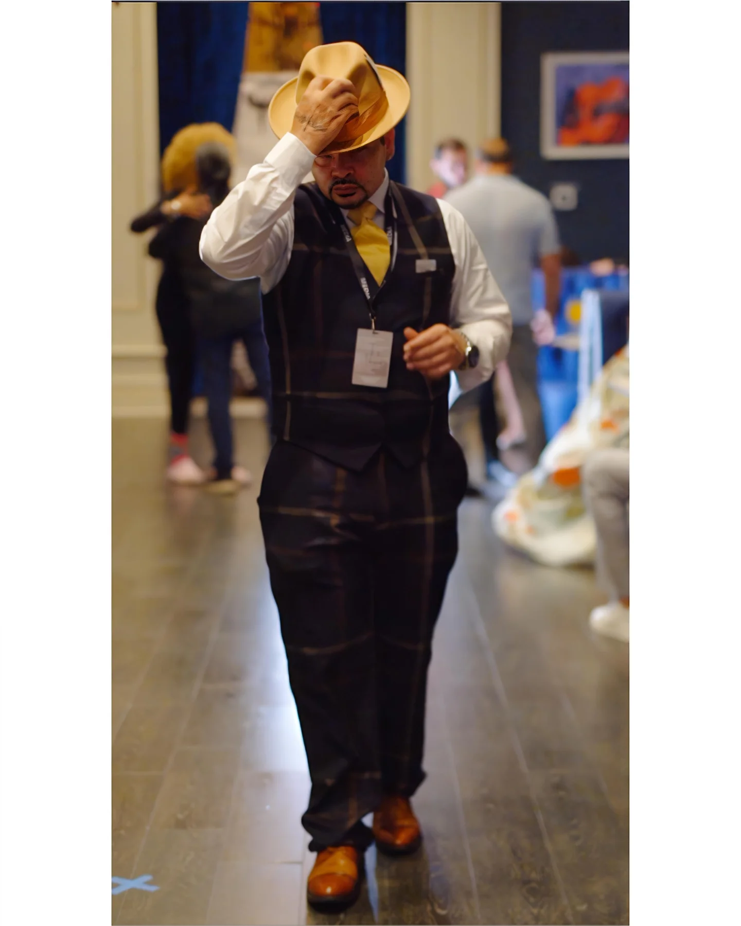 Man in vest and hat walking indoors, holding hat, with blurred people in background.