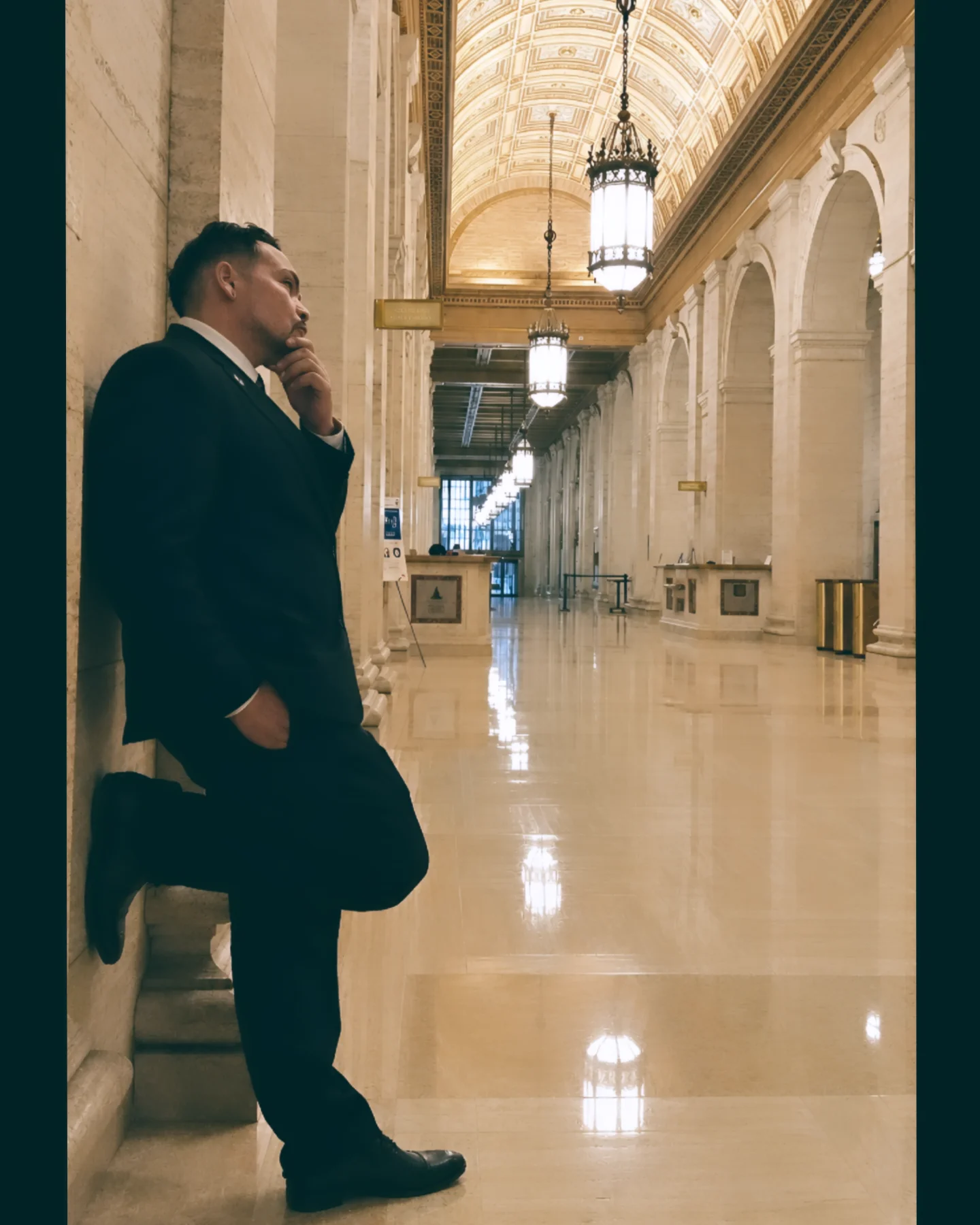 Man in suit leaning against wall in grand hallway with high arched ceiling and hanging lights.