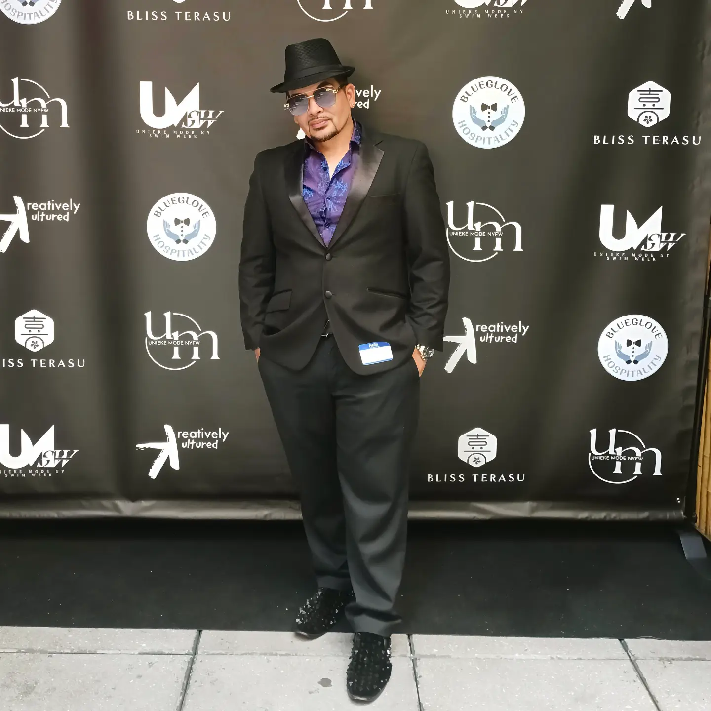 Person in black suit, purple shirt, black hat, glasses, standing on sidewalk in front of black backdrop with logos.