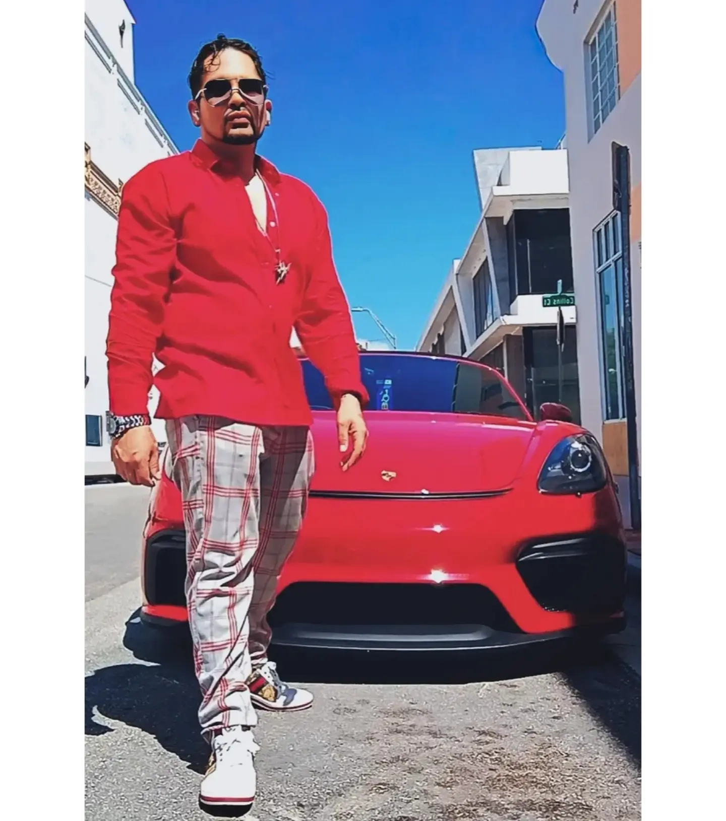 Man in red jacket and plaid pants standing next to a red sports car on a city street under a blue sky.