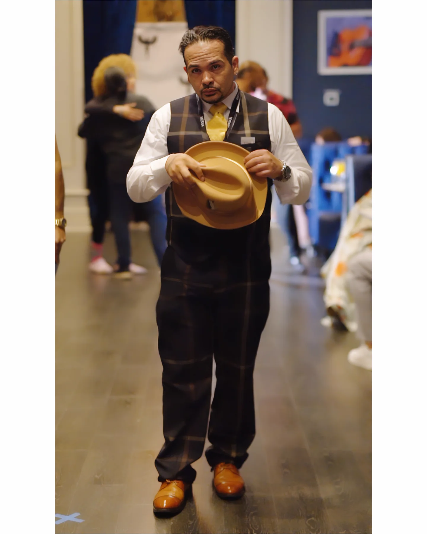 Man holding a hat in a room with people and artwork, standing on a wooden floor.