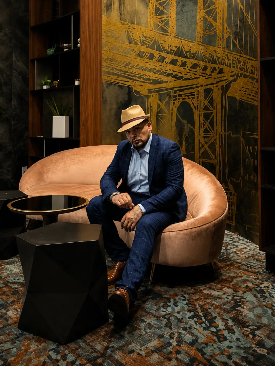 Man in a hat and suit sitting on a pink sofa in a room with a large gold mural and dark wooden shelves.