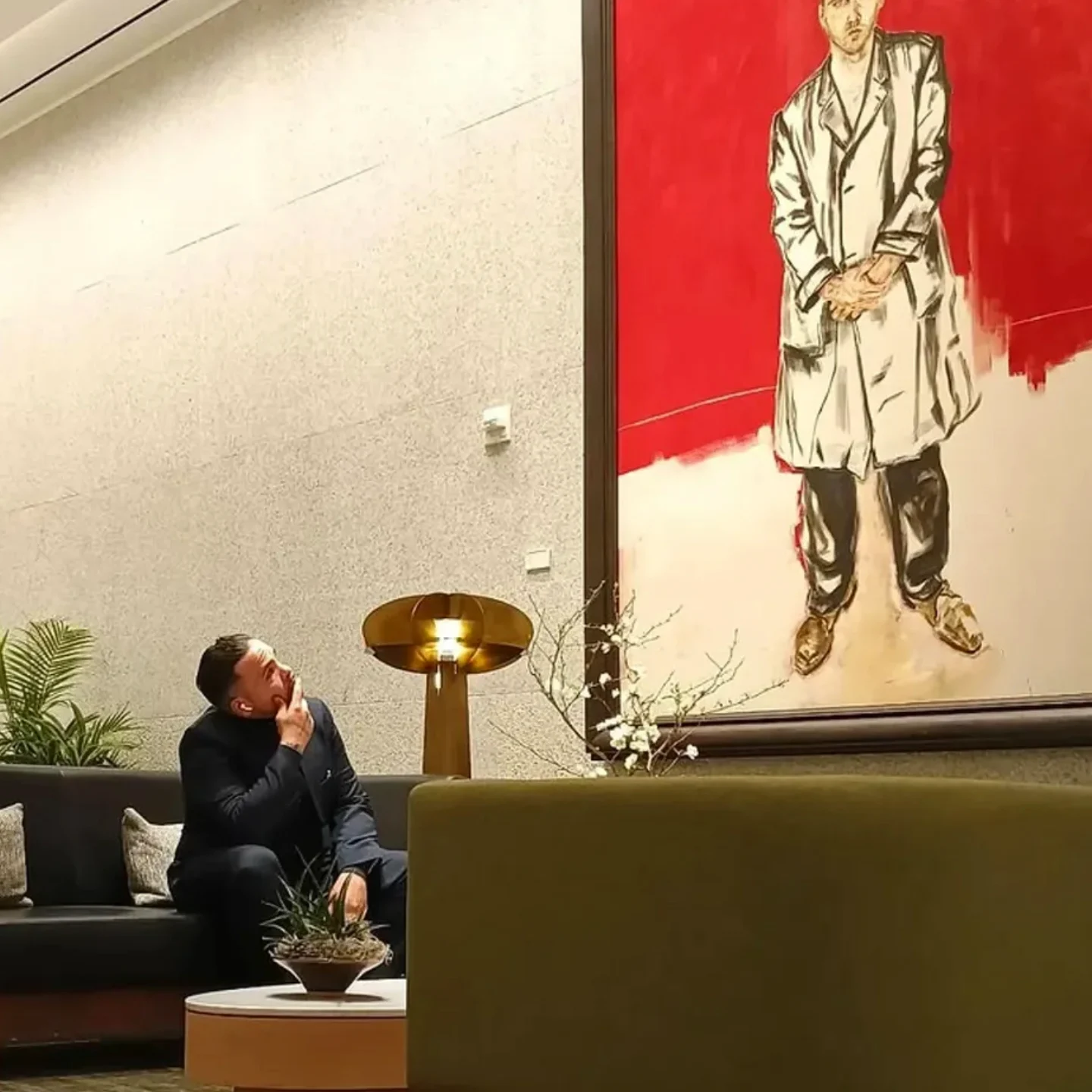 A man sitting on a sofa, looking at a large painting of a person in a coat, in an indoor setting.