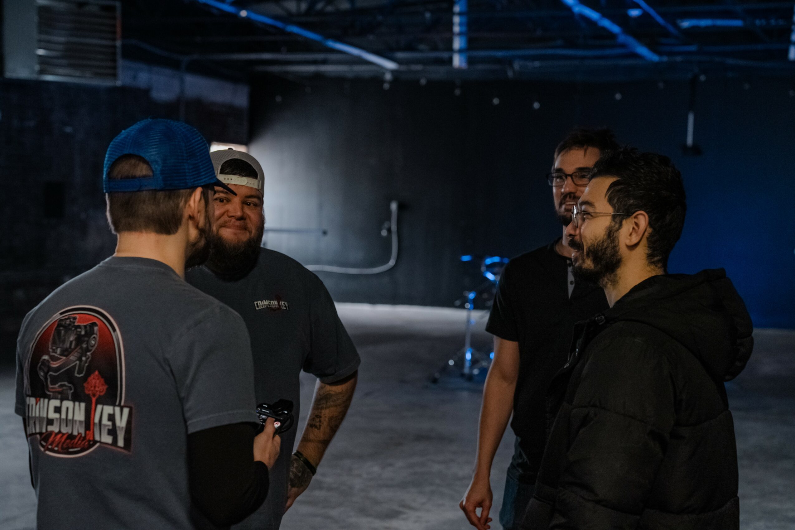 Four men standing and talking in a dimly lit indoor space with a concrete floor.