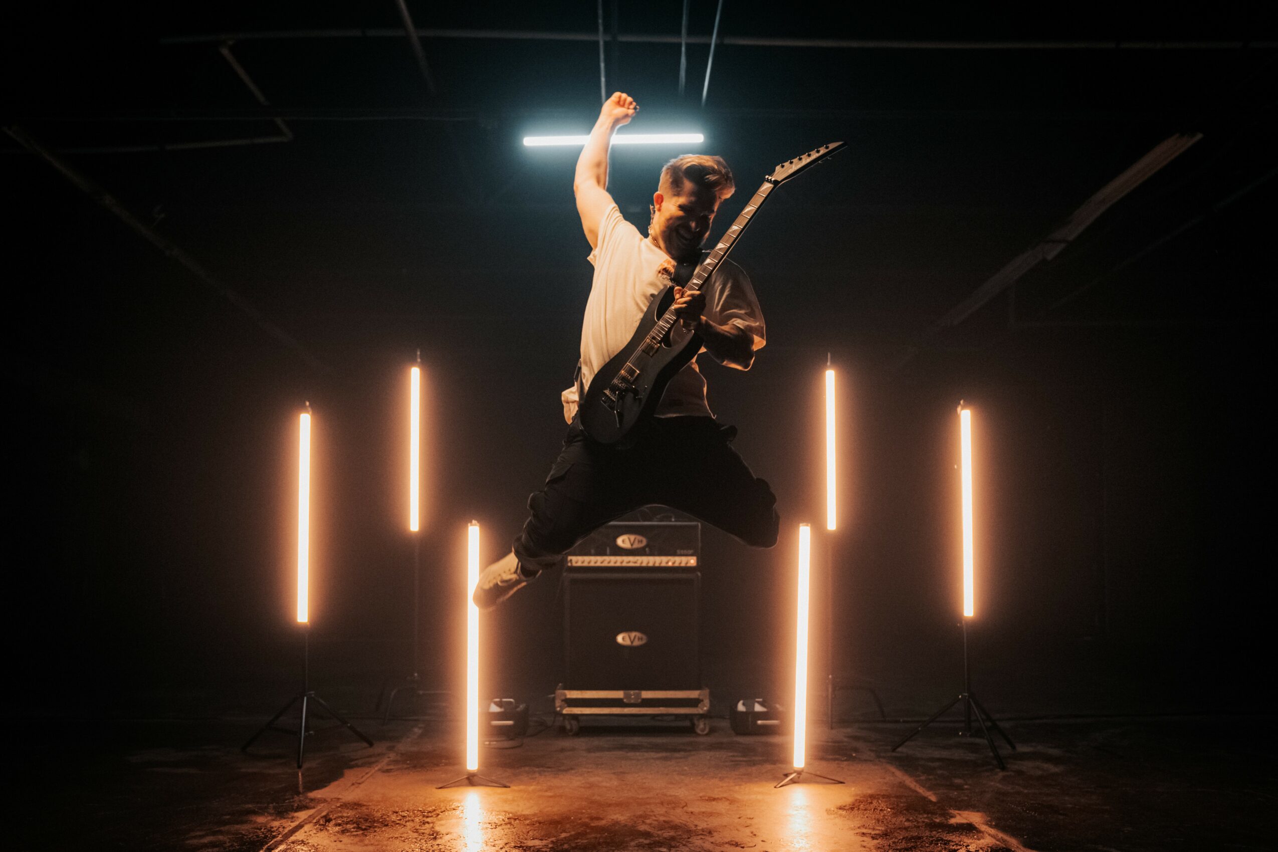 Person jumping with guitar on stage, illuminated by vertical lights, in a dark setting.