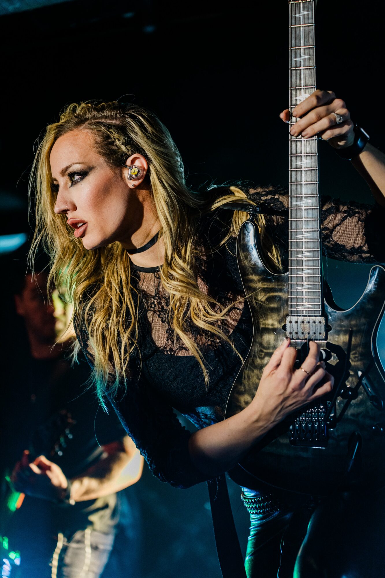 Woman with long blonde hair holding an electric guitar on stage, wearing black clothing and makeup, focused expression.