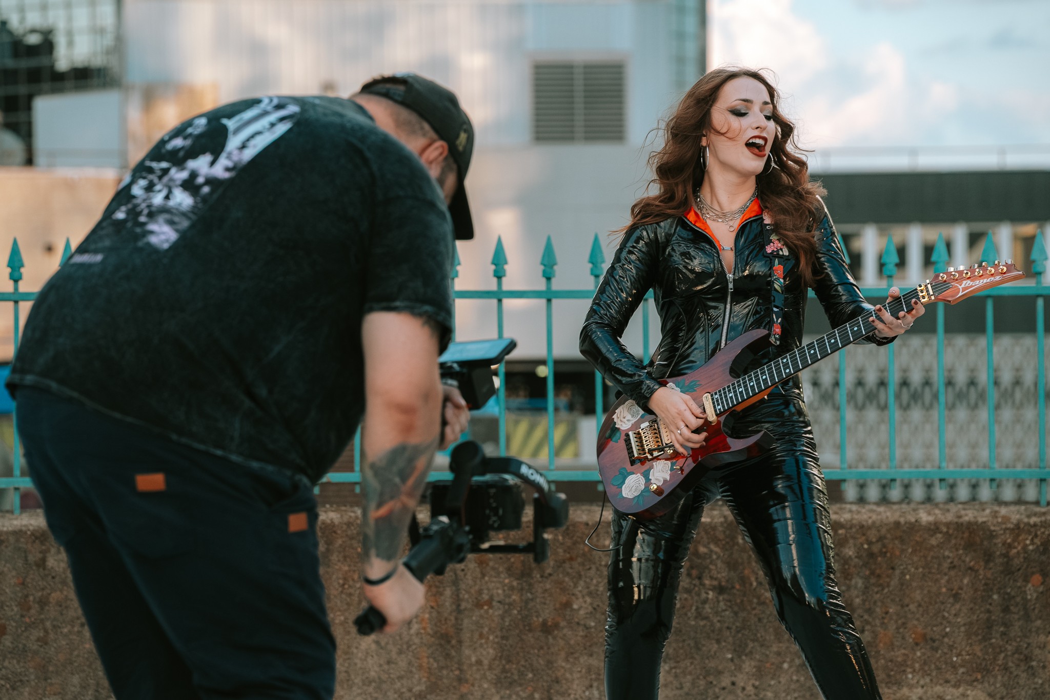A woman with long hair wearing a black shiny outfit playing an electric guitar outdoors, with a person filming her.
