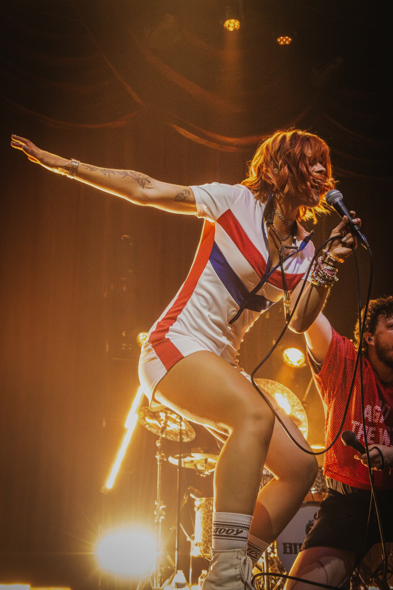 Female singer with red hair performs on stage, holding a microphone, with one arm extended, wearing a striped outfit.