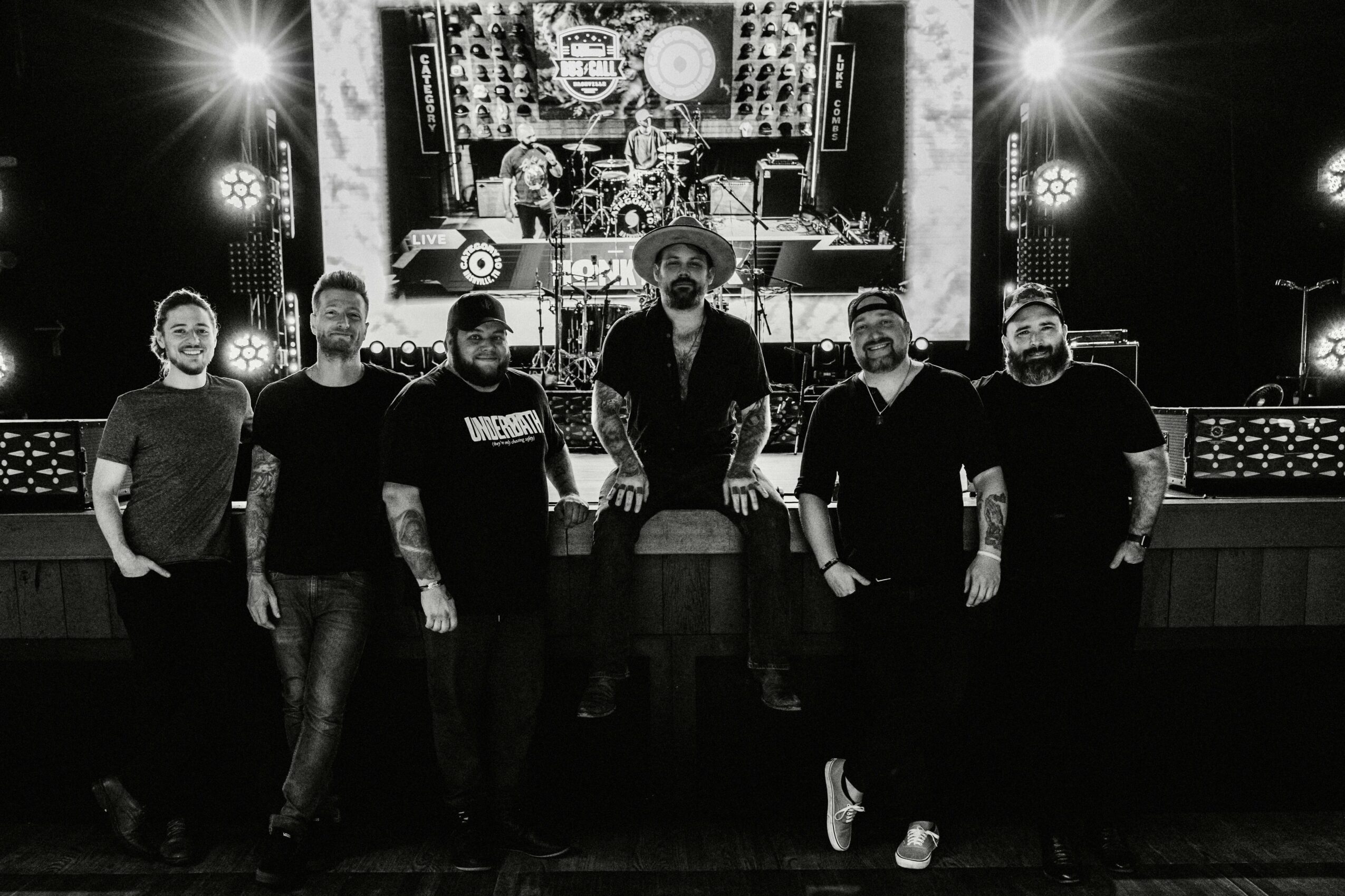 Six people standing on stage in front of a large screen with stage lights, in black and white.