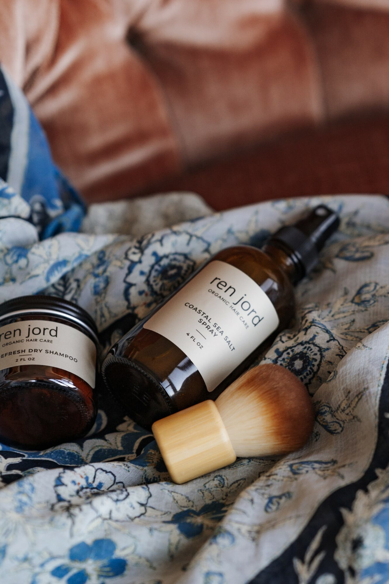 Skincare products and a brush on patterned fabric with a brown sofa in background.