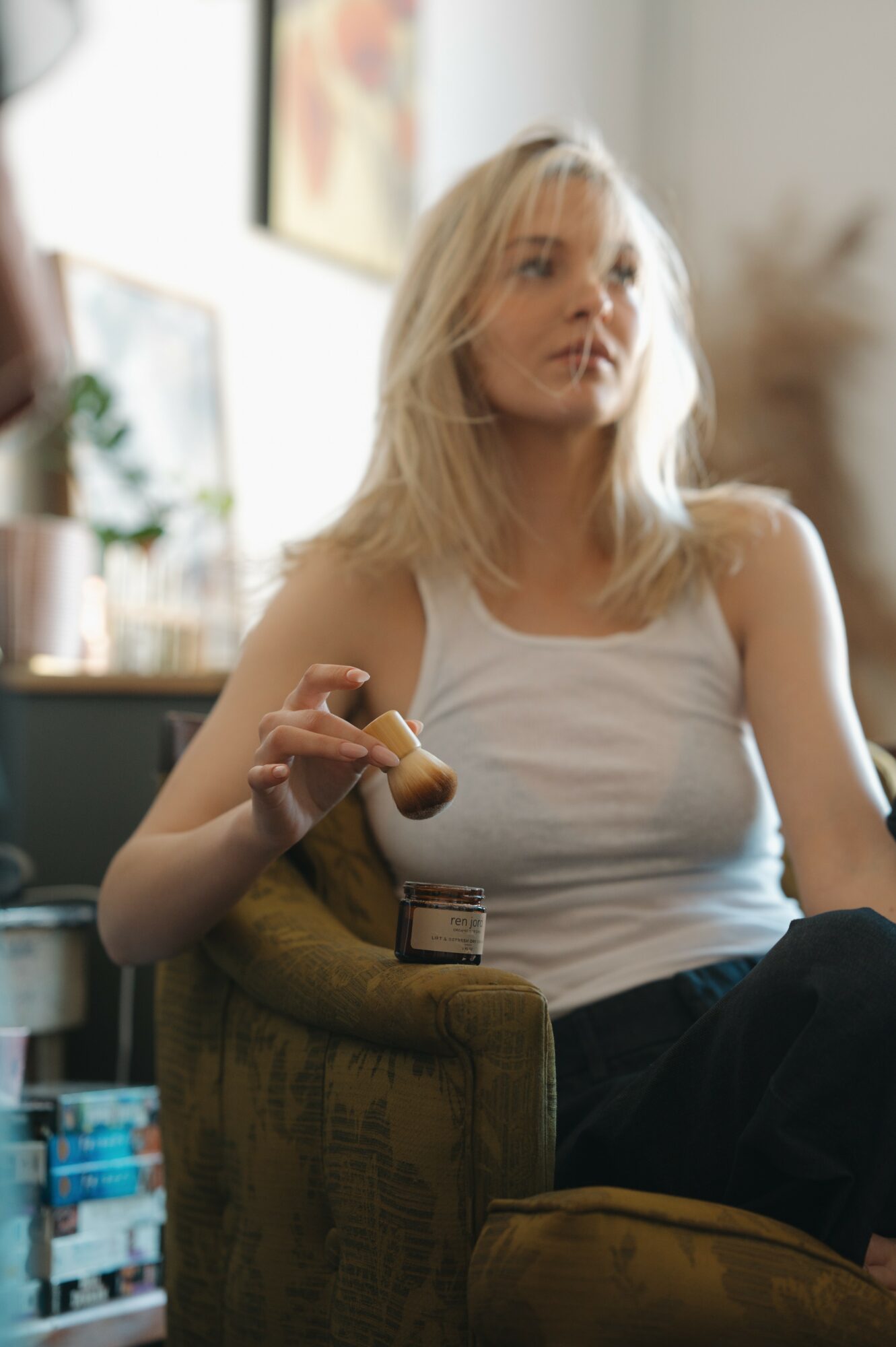 Young woman with blonde hair sitting on a green armchair, holding a pipe, in a room with artwork and plants.