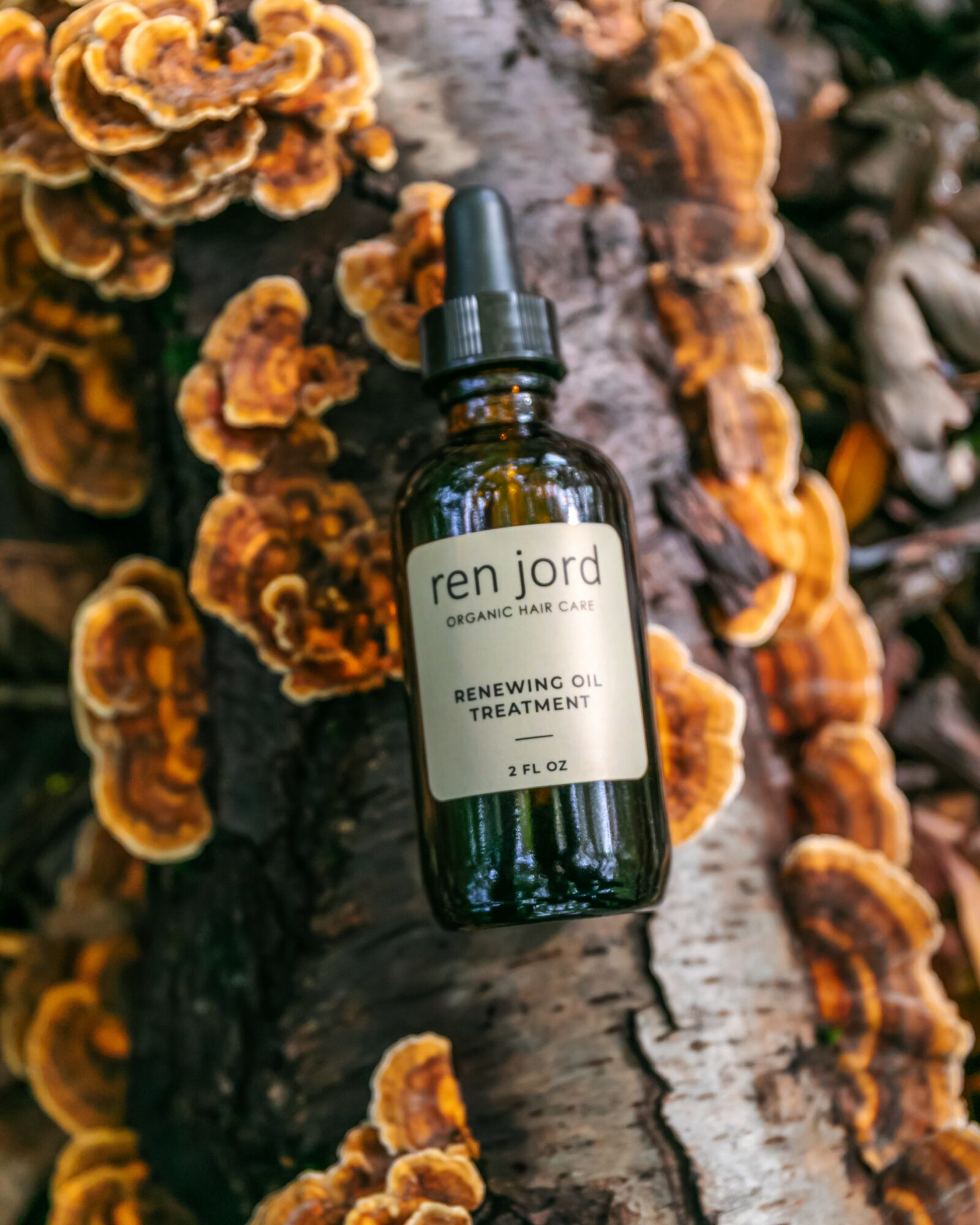A dark green glass bottle with a dropper cap, labeled 'ren jord' and 'Renewing Oil Treatment,' placed on a log with orange fungi.