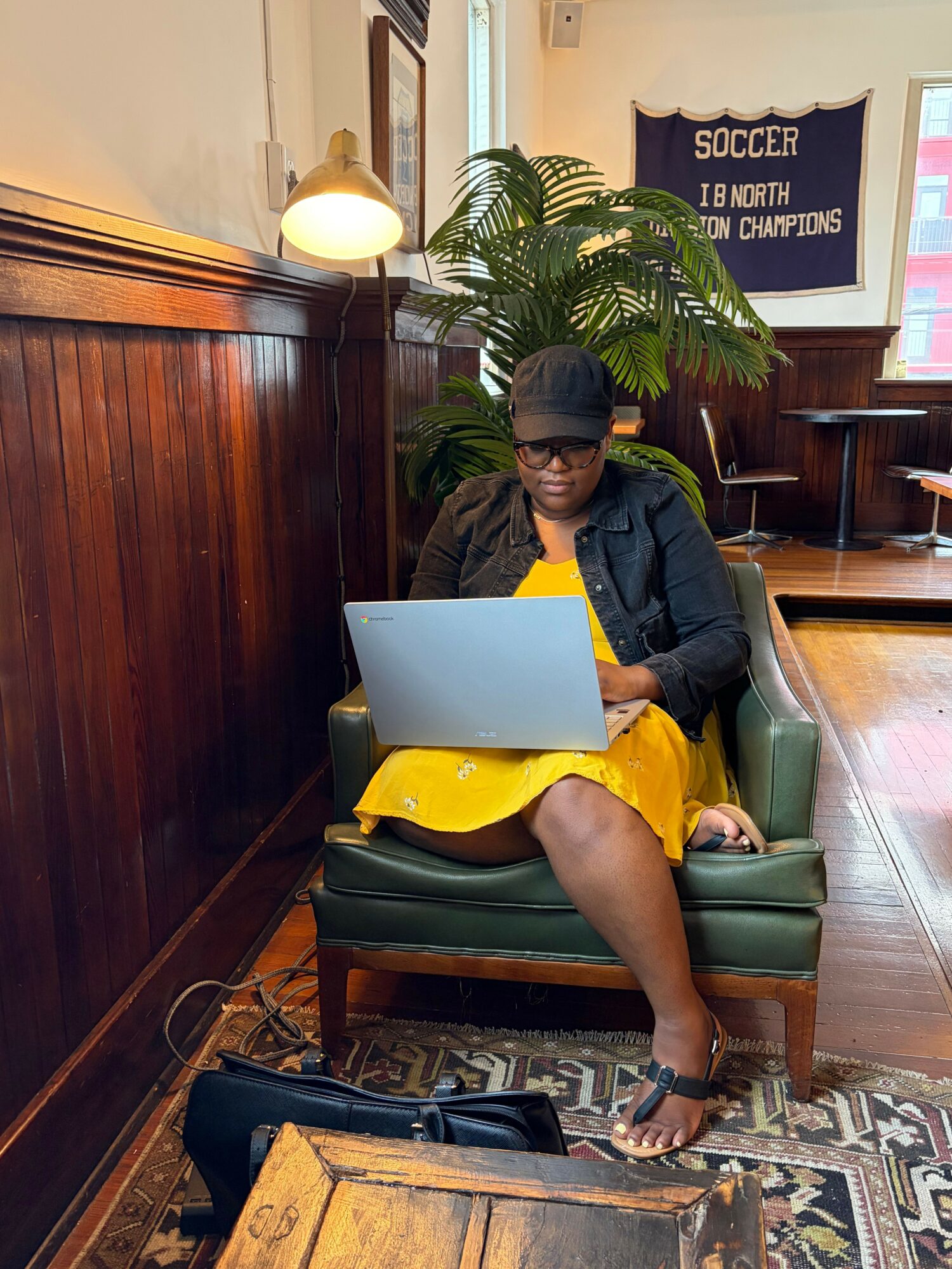 Person sitting on a green armchair using a laptop, with a large plant and wooden furniture in the background.
