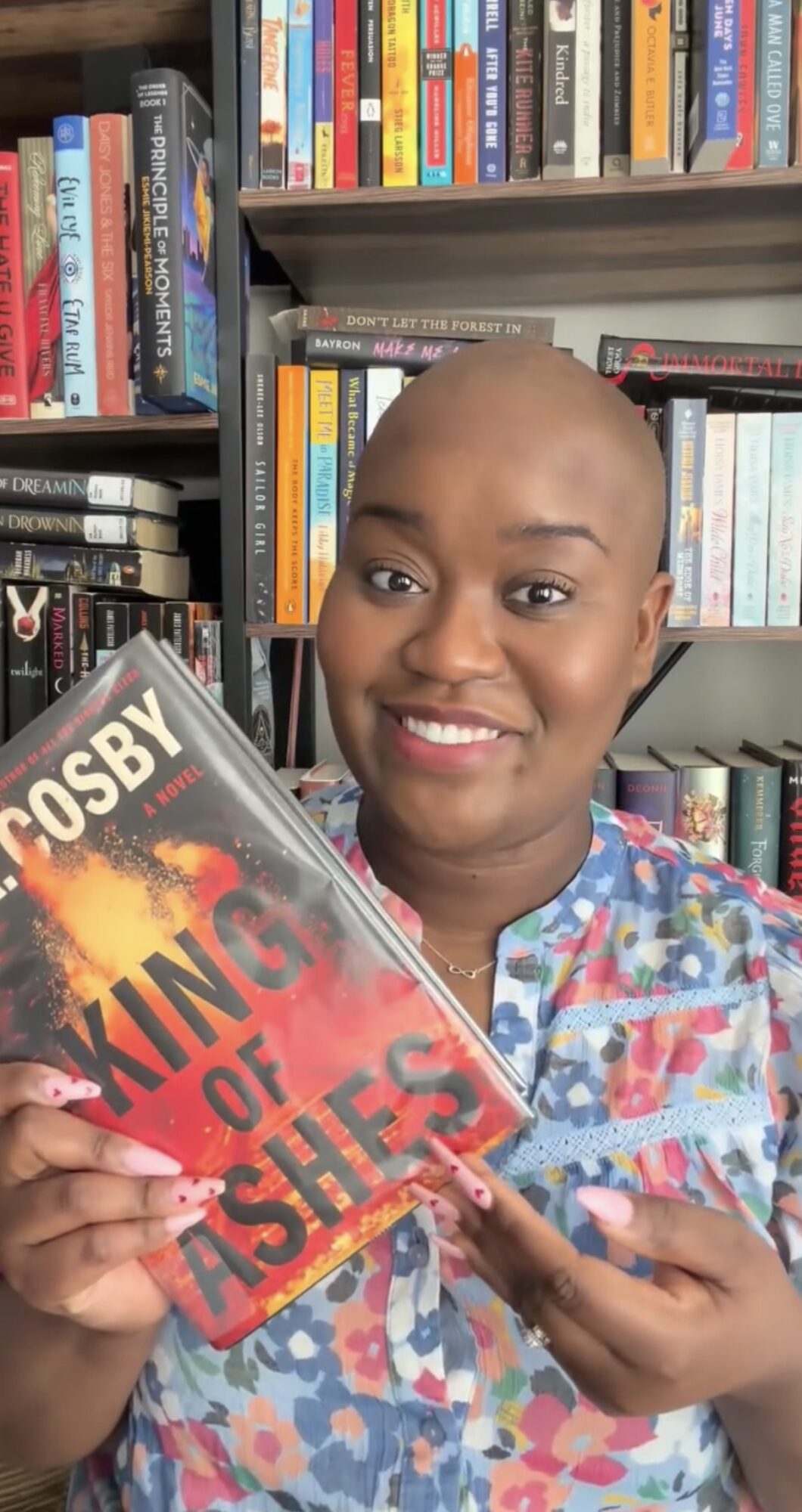 Person with a shaved head smiling and holding a book titled 'King of Ashes' in front of a bookshelf.