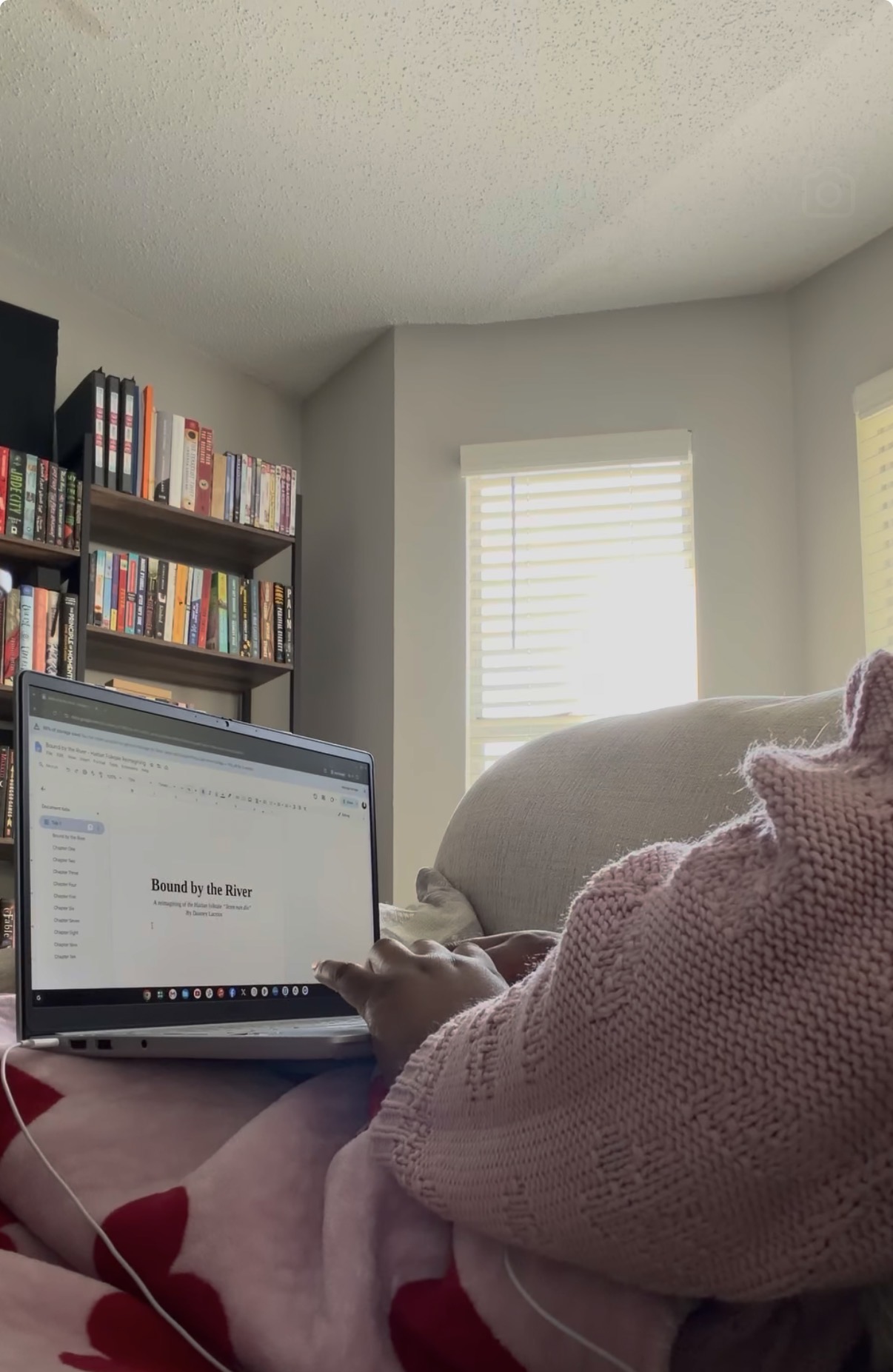 Person using a laptop on their lap in a room with a bookshelf and window, wearing a pink sweater.