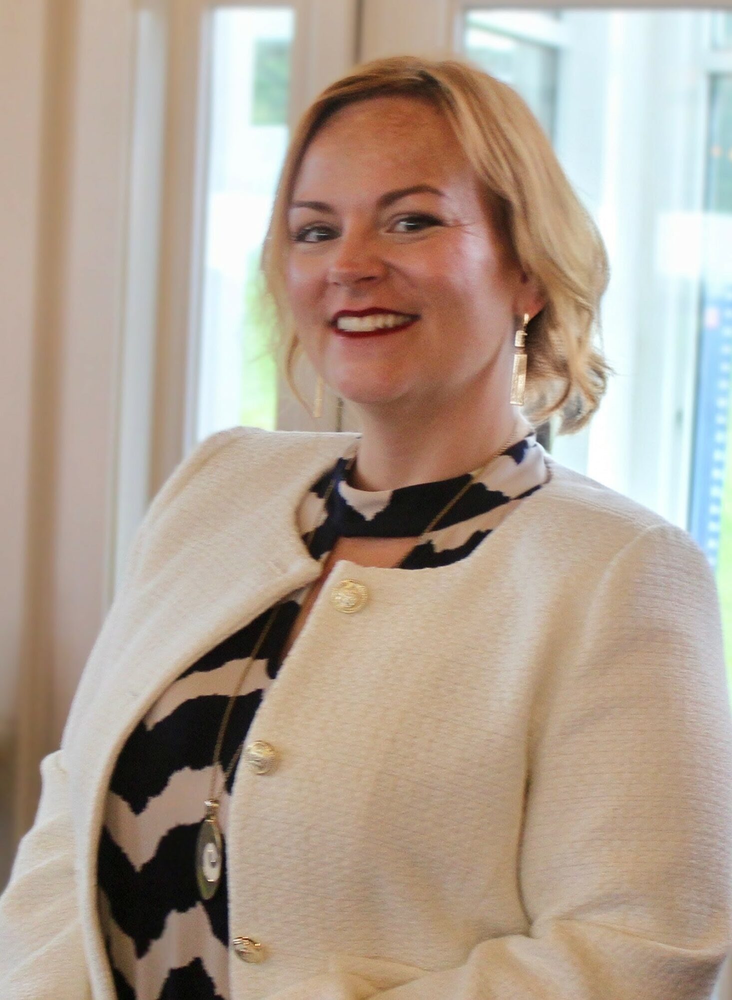 Smiling woman with short blonde hair wearing a cream jacket and patterned blouse, standing indoors near windows.