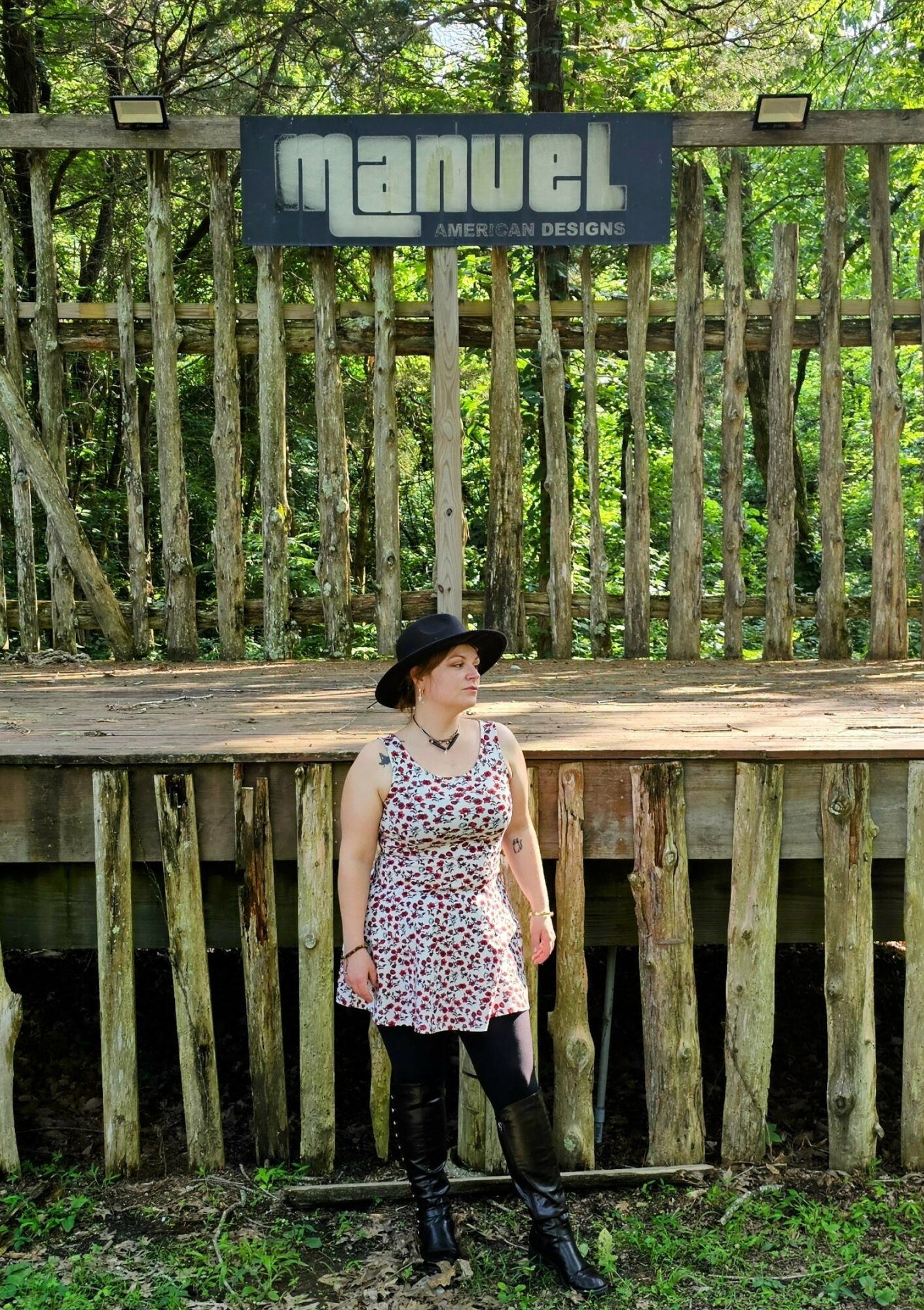Woman in a floral dress and black hat standing in front of a wooden fence with trees behind.