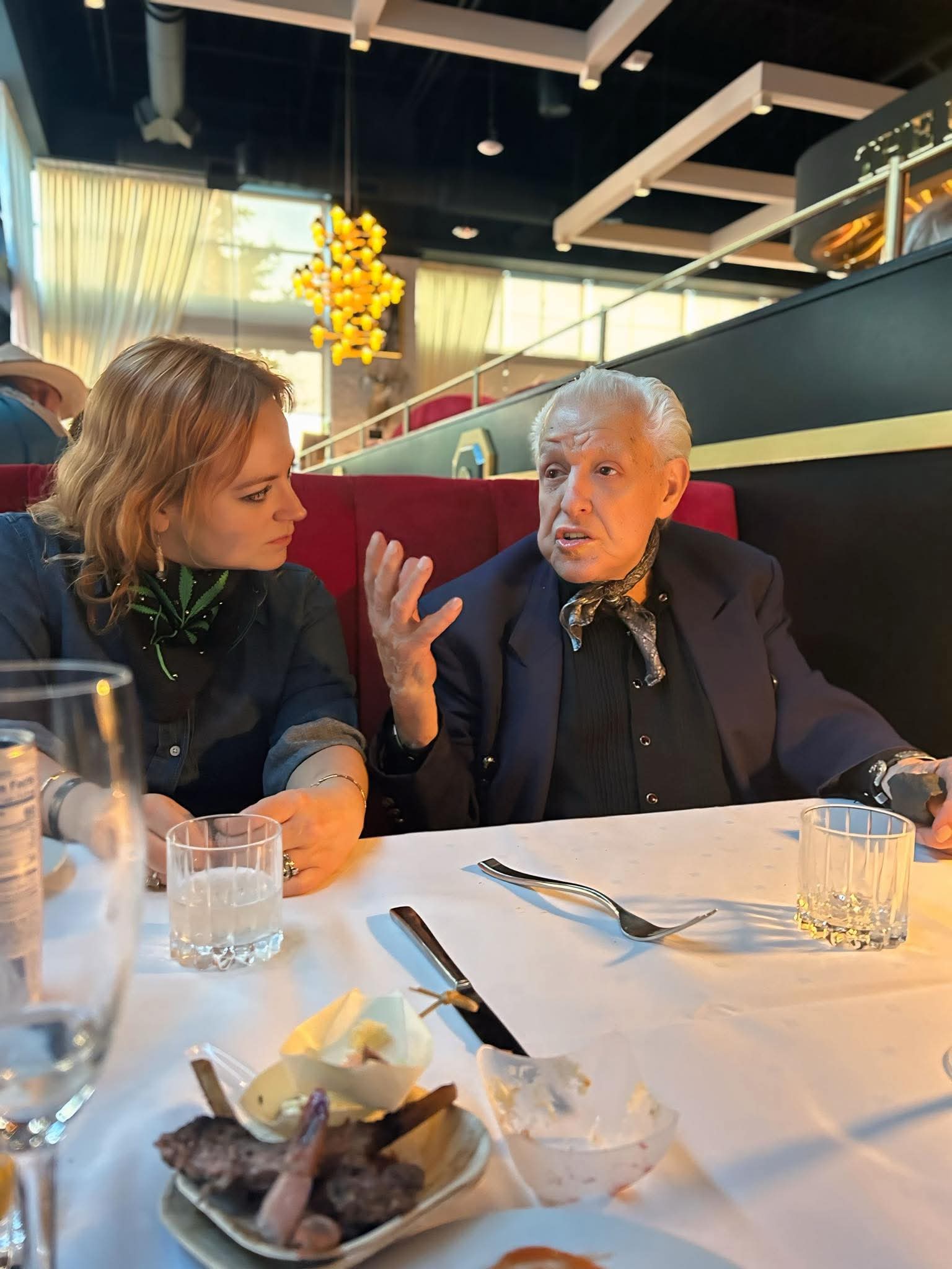Two women sit at a table in a restaurant, engaged in conversation. The table has glasses, utensils, and a dish with food.