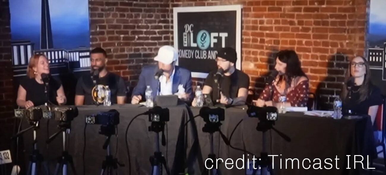 Six people sit at a panel table on a stage with a brick wall and a sign behind them, with microphones in front.