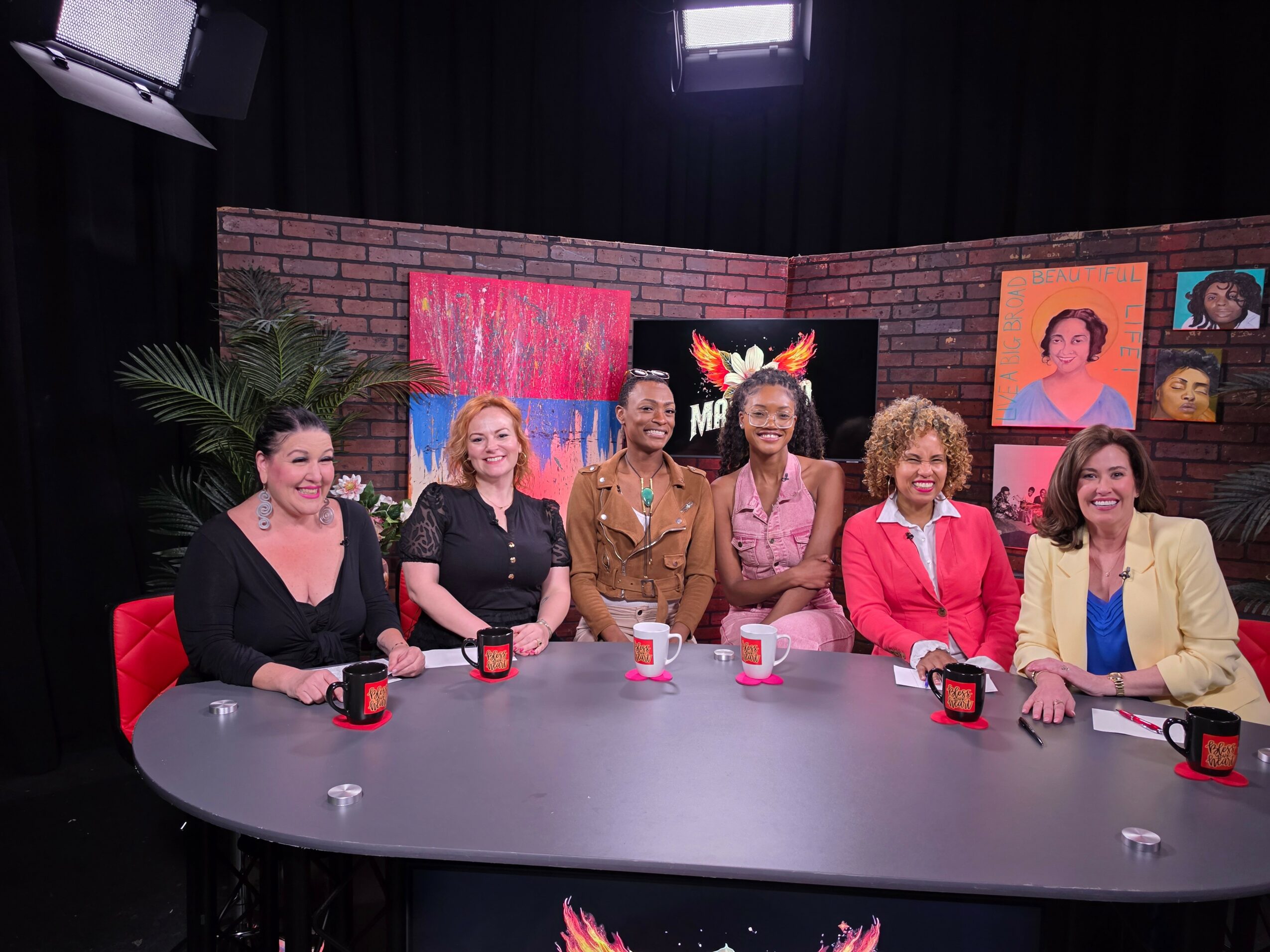 Six women sitting at a curved table with mugs, in a studio with colorful artwork and a brick wall background.
