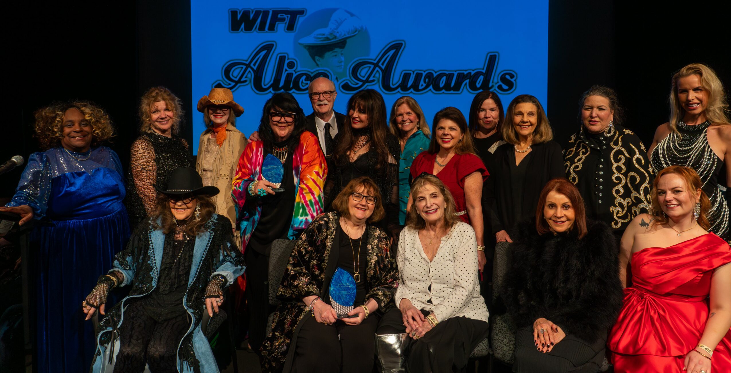 Group of people on stage at an awards event, with a blue background and text, some smiling and dressed formally.