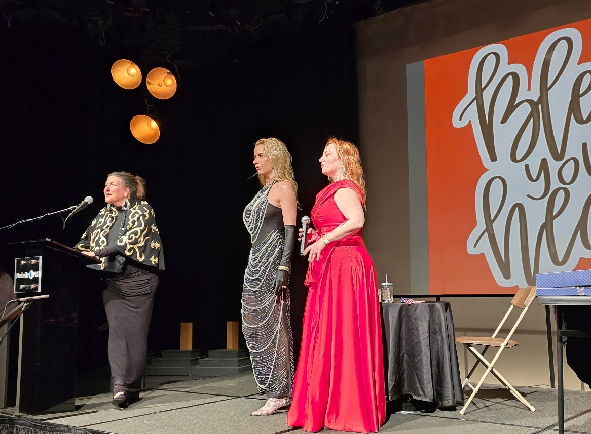 Three women on stage, one speaking at a microphone, two standing nearby, with a large screen displaying text behind them.