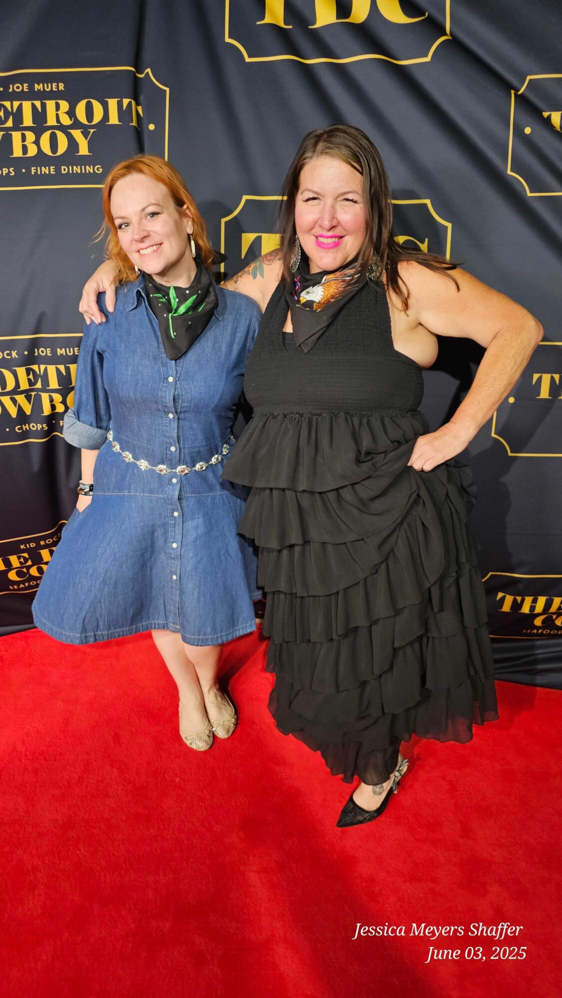 Two women in formal dresses standing on a red carpet, smiling, with a black backdrop behind them.