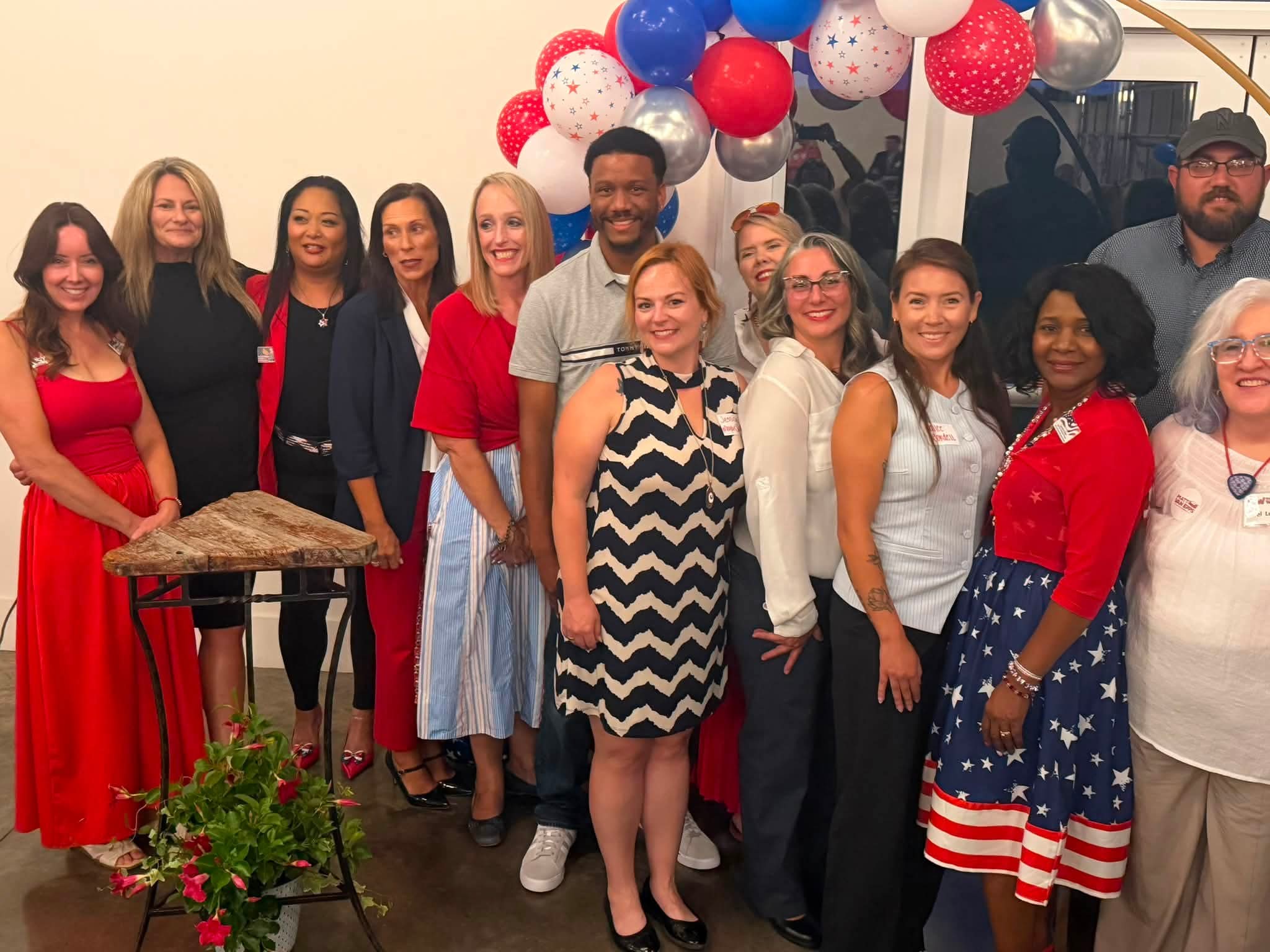Group of people standing indoors with balloons and decorations, smiling for a photo, some wearing red, white, and blue clothing.