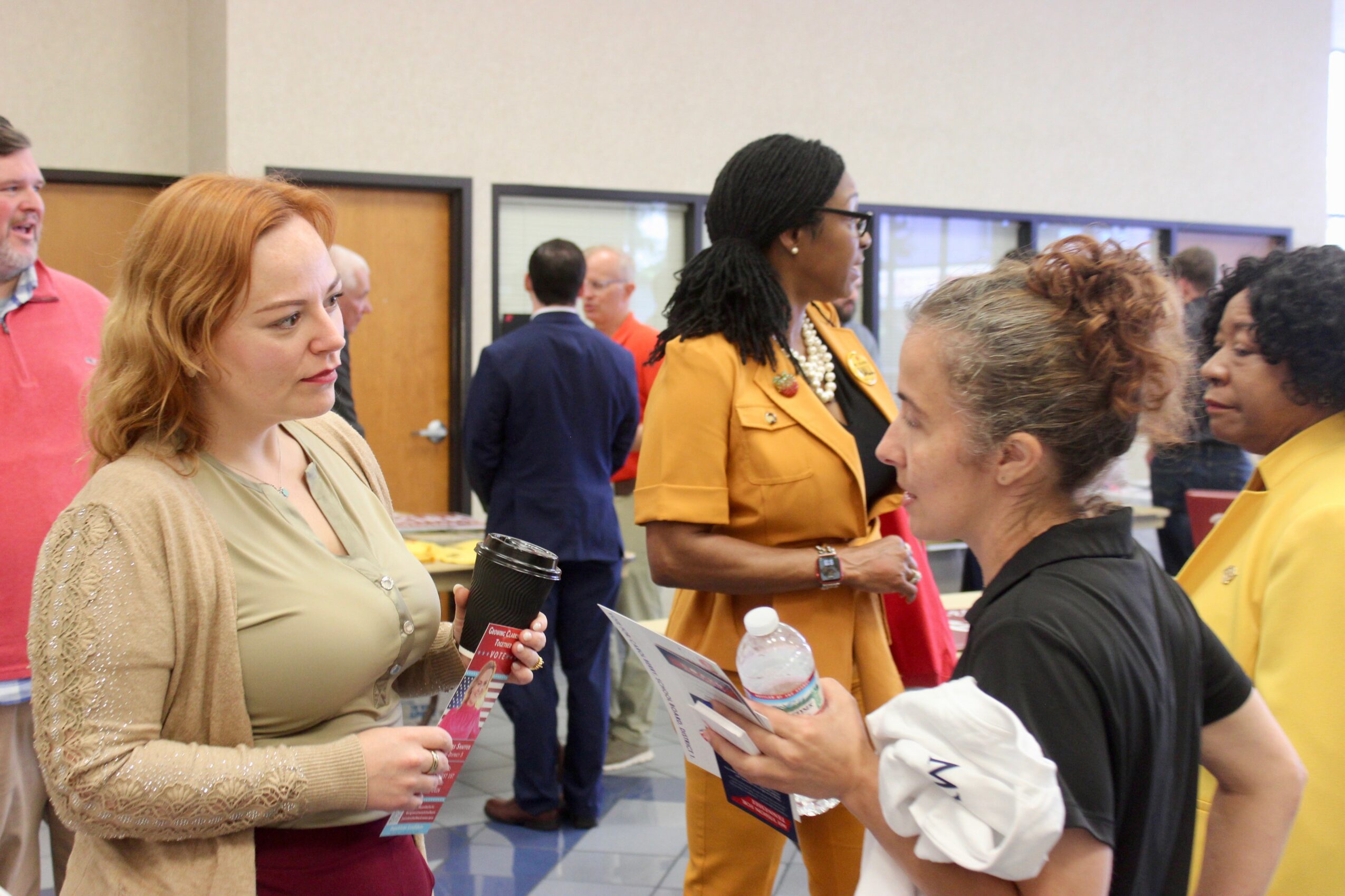 Group of women engaged in conversation indoors, some holding papers and water bottles, with a background of people and screens.