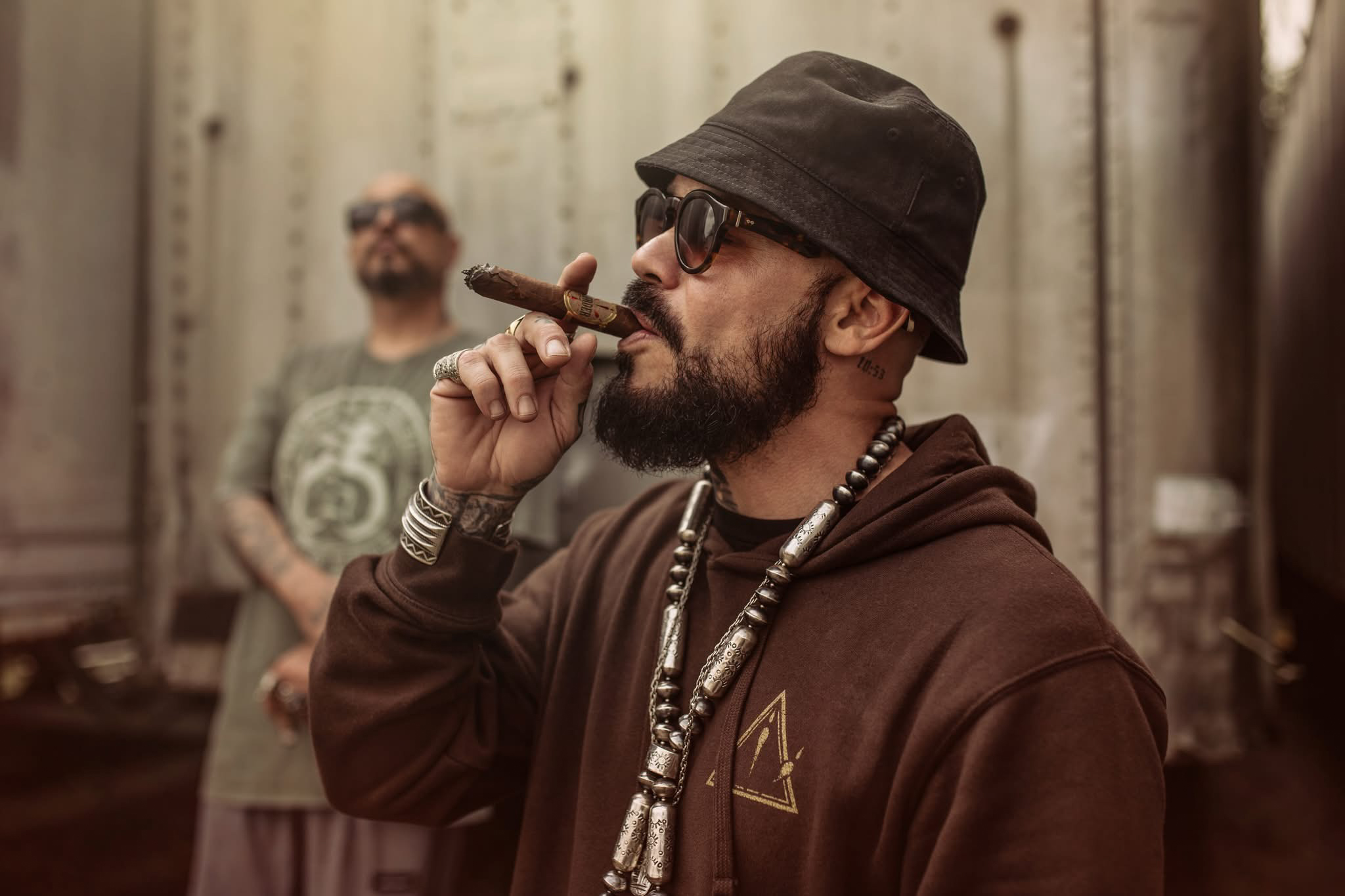 Man with glasses, hat, and beard smokes a cigar in an industrial setting, with another person blurred in the background.